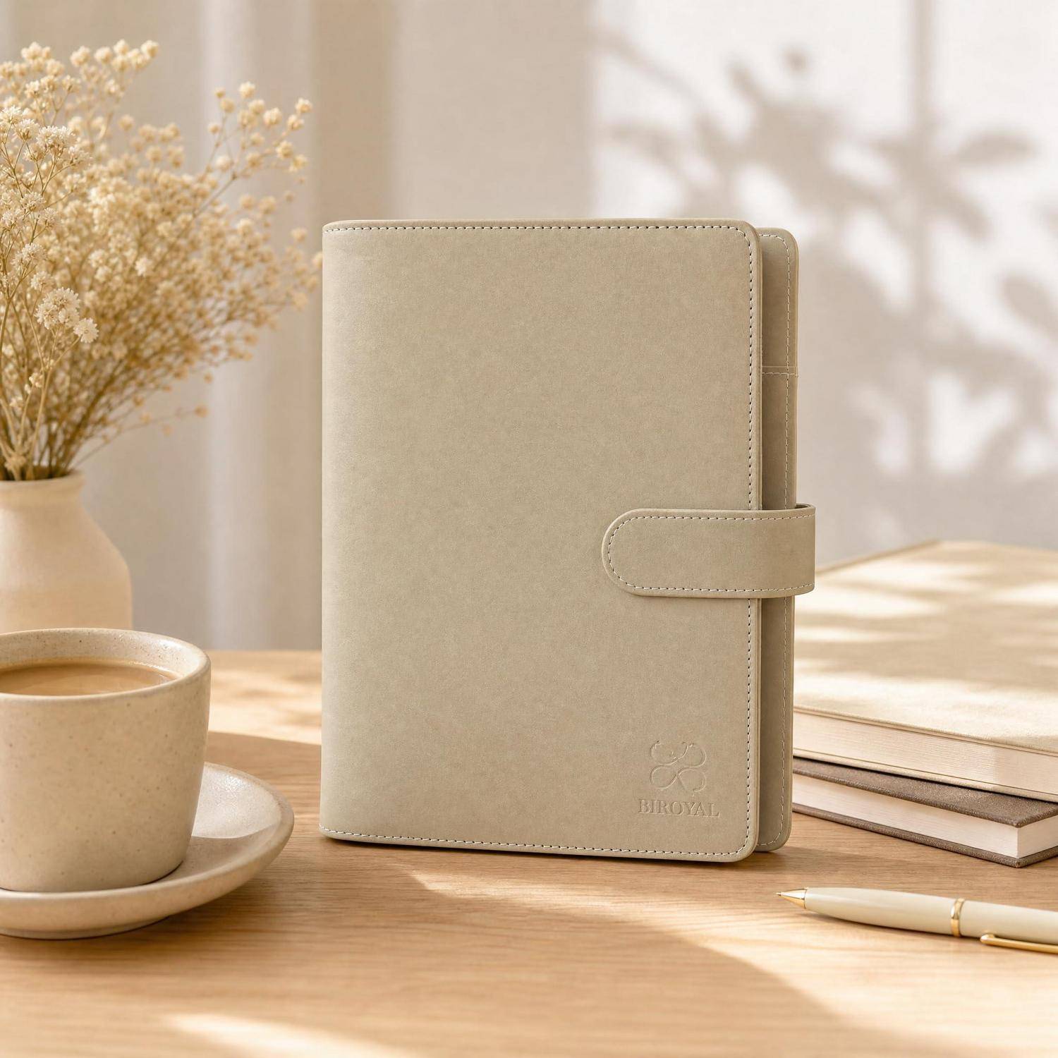 Beiger Lederplaner oder ein Tagebuch, das auf einem Holzschreibtisch neben einer Kaffeetasse, einer Vase mit getrockneten Blumen, gestapelten Büchern und einem Stift im warmen Licht steht.