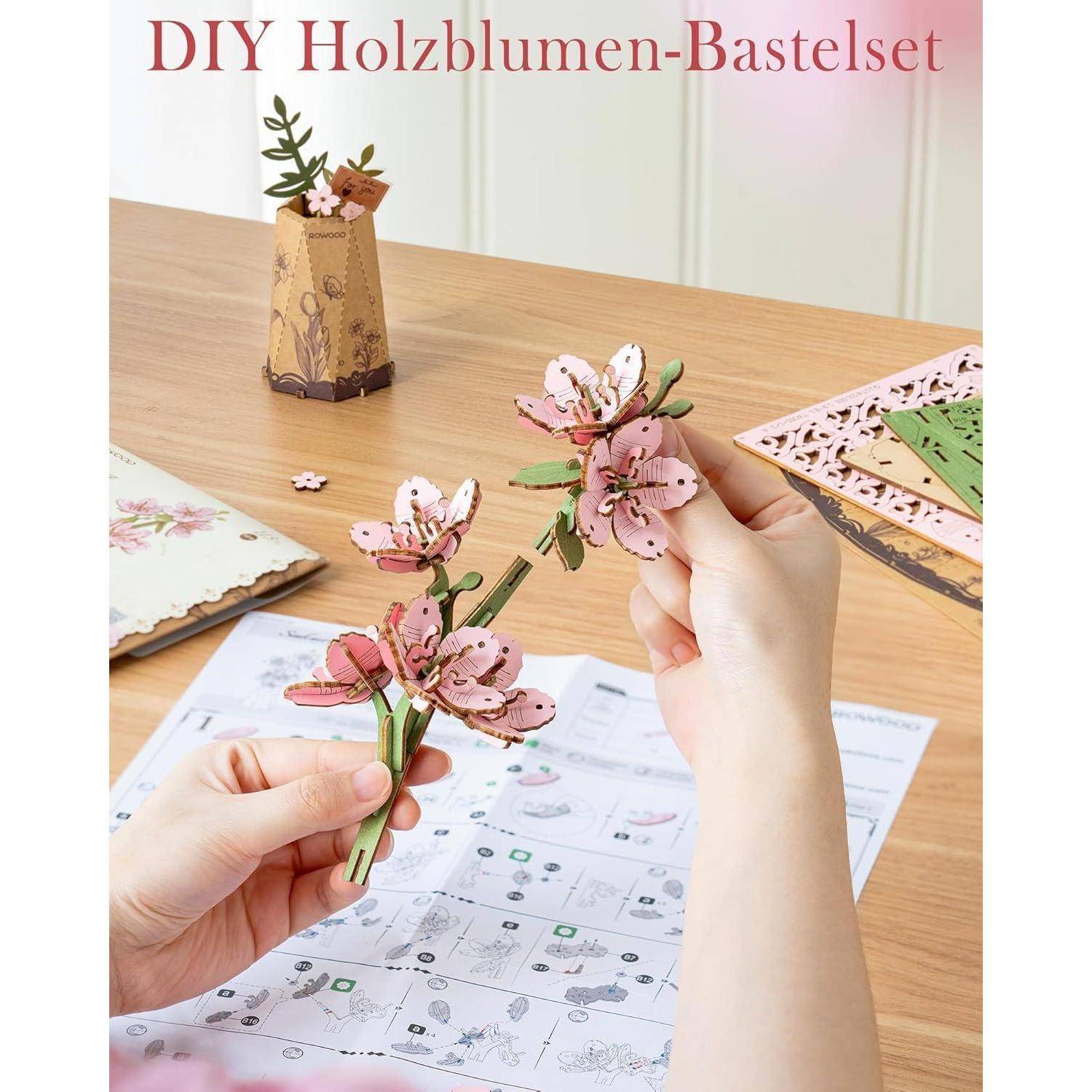 Hände, die rosa Holzblumen aus einem DIY-Bastelset auf einem Tisch zusammensetzen, mit fertigen Blütenteilen und dem deutschen Titel „DIY Holzblumen-Bastelset