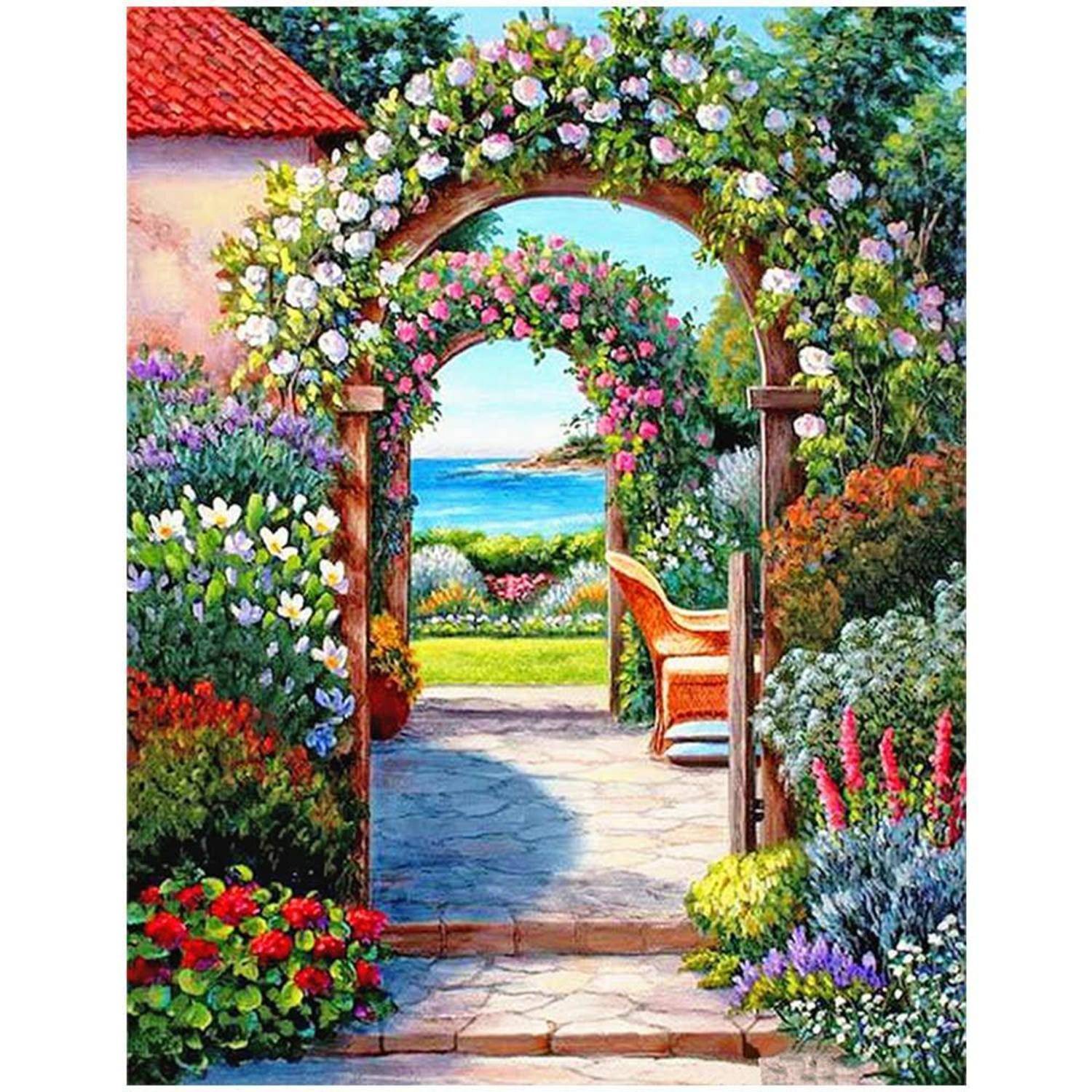 Mit Blüten bedeckte Gartenbögen öffnen sich zu einem Steinweg, mit einem Korbstuhl und einem Blick auf das blaue Meer jenseits bunter blühender Beete.