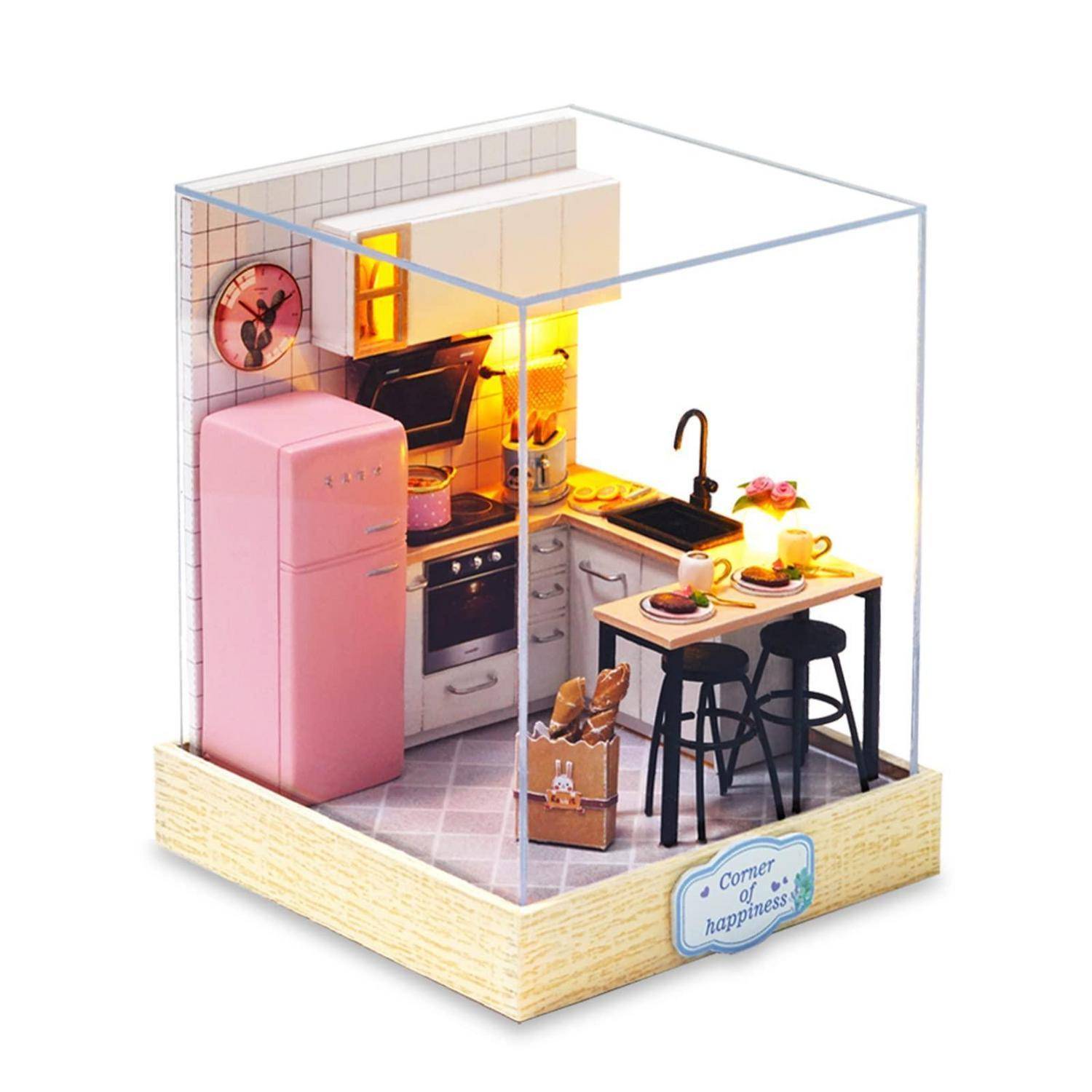 Eine miniaturisierte offene Küchenlandschaft mit einem rosa Retro-Kühlschrank, Herd, Spüle, Tisch mit Hockern, warmer Beleuchtung und einem Schild mit der Aufschrift „Ecke des Glücks