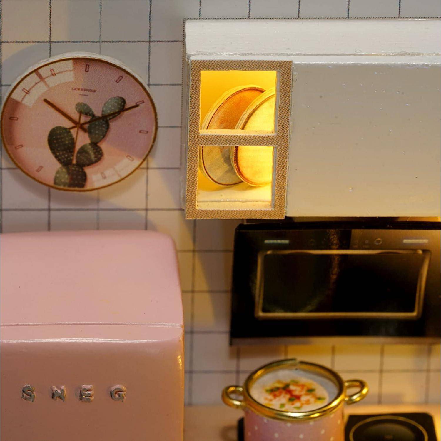 Miniaturküchen-Szene mit einem rosa SMEG-Kühlschrank im Stil, einer Wanduhr, einem Herd, einem beleuchteten Schrank und einem kleinen Topf auf der Kochplatte.