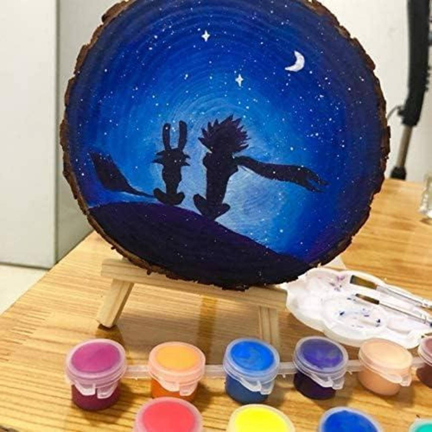 Bemalte Holzscheibe mit silhouettierten Kaninchen und Igel unter einem sternenklaren Mondhimmel, mit Farbdosen und Pinseln darunter