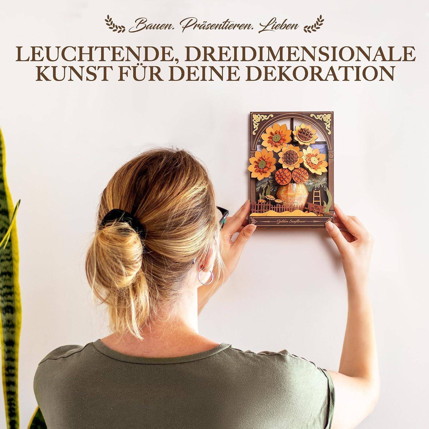 Eine Frau hängt ein dreidimensionales Blumen-Wandkunstwerk auf; deutscher Text lautet „LEUCHTENDE, DREIDIMENSIONALE KUNST FÜR DEINE DEKORATION