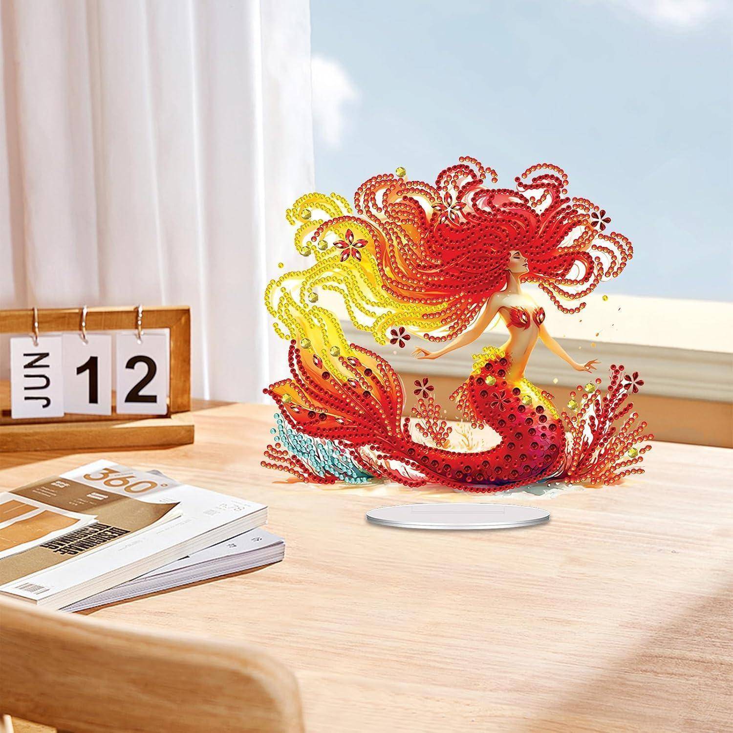 Dekorative Meerjungfrauenfigur mit fließenden roten und gelben Wirbeln auf einem Schreibtisch neben einem Kalender vom 12. Juni und Zeitschriften, in der Nähe eines Fensters.