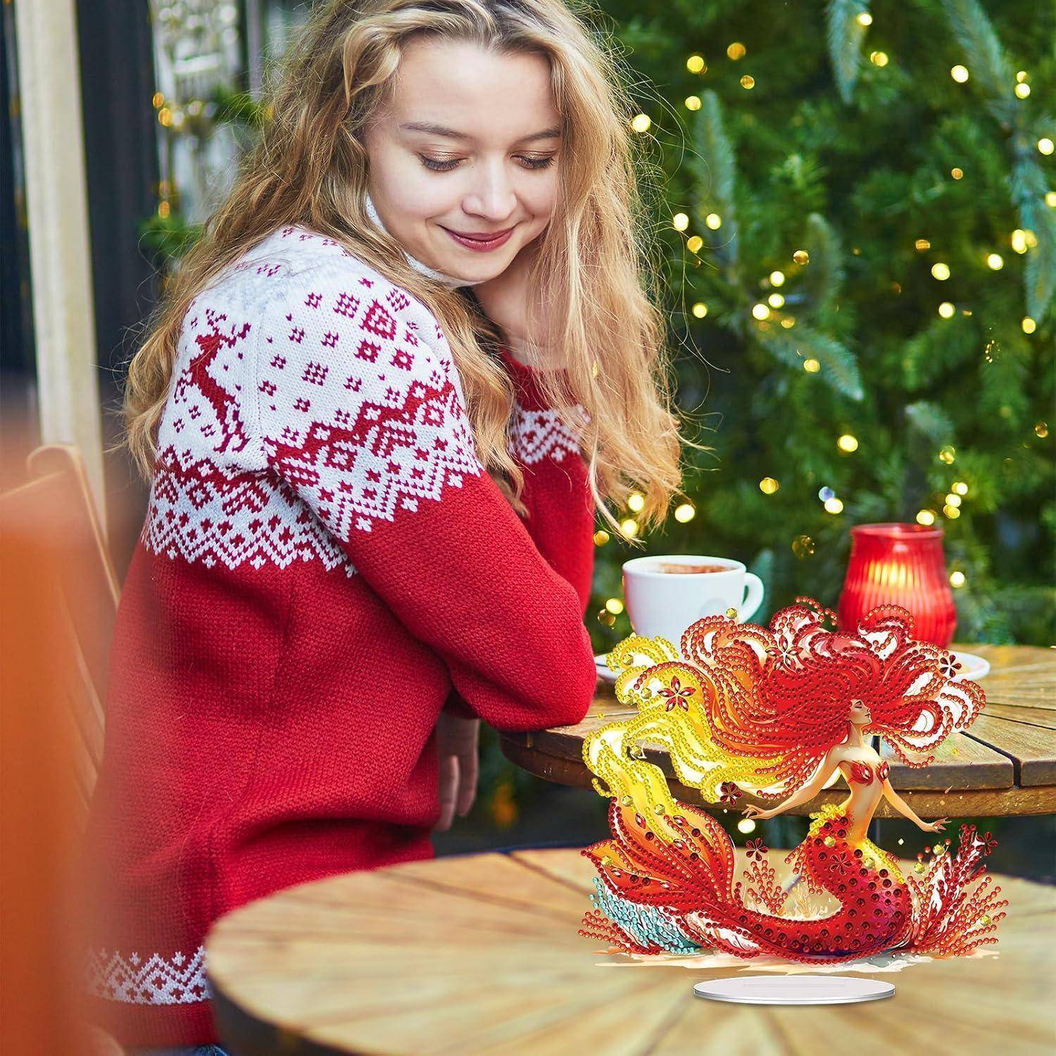 Eine junge Frau in einem roten Weihnachtspullover sitzt neben einem geschmückten Weihnachtsbaum und schaut sich eine bunte Hahnen-Weihnachtskugel auf einem Tisch an, neben einer Tasse und einer Kerze.