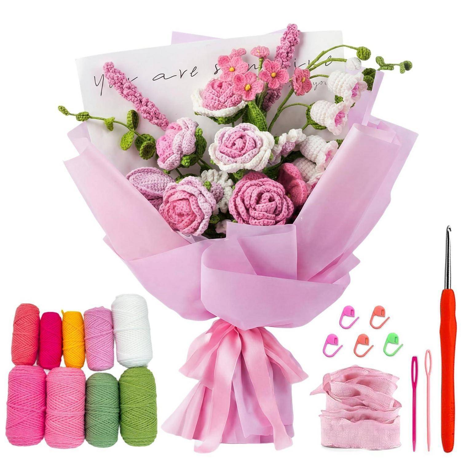 Gehäkelter rosa und weißer Blumenstrauß, eingewickelt in rosa Papier, mit Wollknäueln und Häkelhaken, die als verwendete Materialien zum Herstellen gezeigt werden.