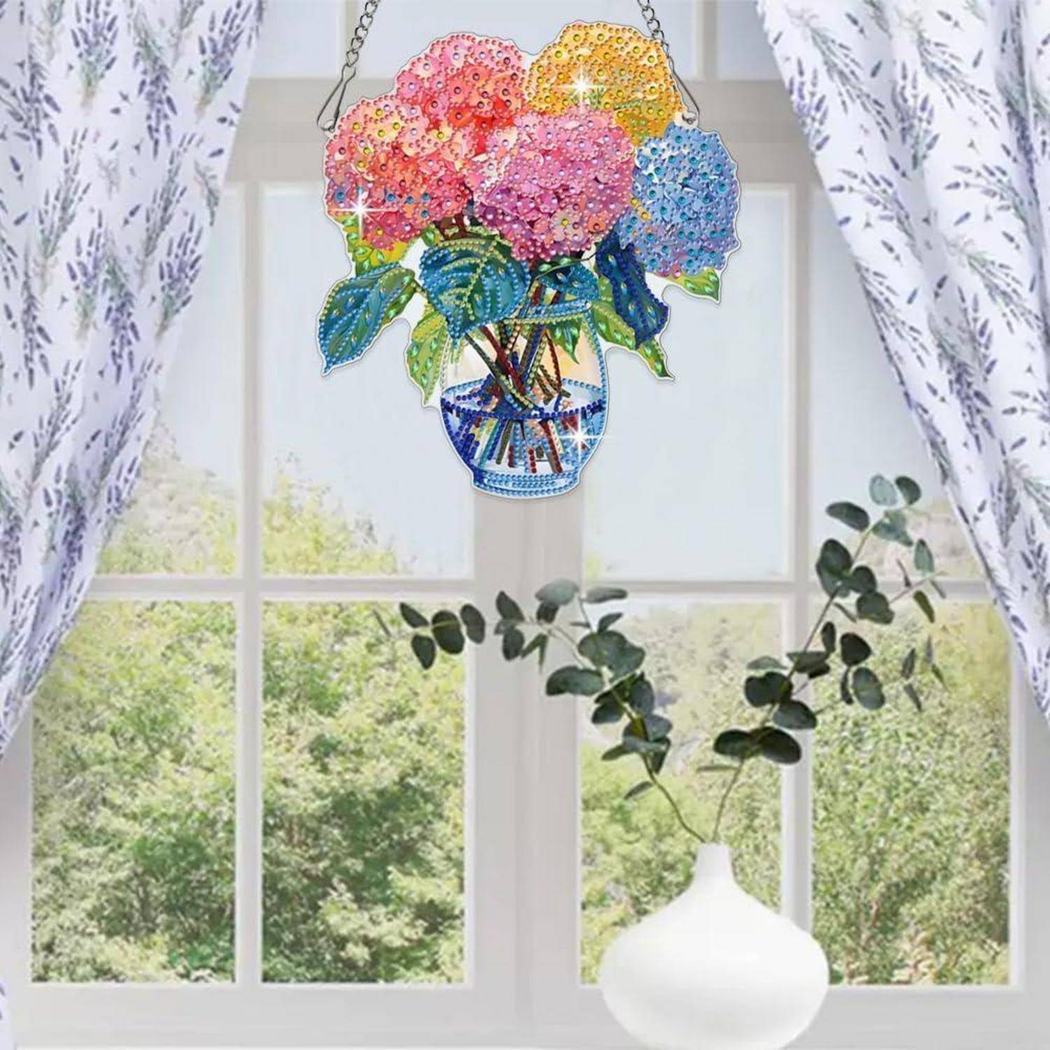 Bunter Blumenschmuck aus Perlen in einer Vase hängt vor einem sonnendurchfluteten Fenster mit weißen Vorhängen und Grünflächen draußen.