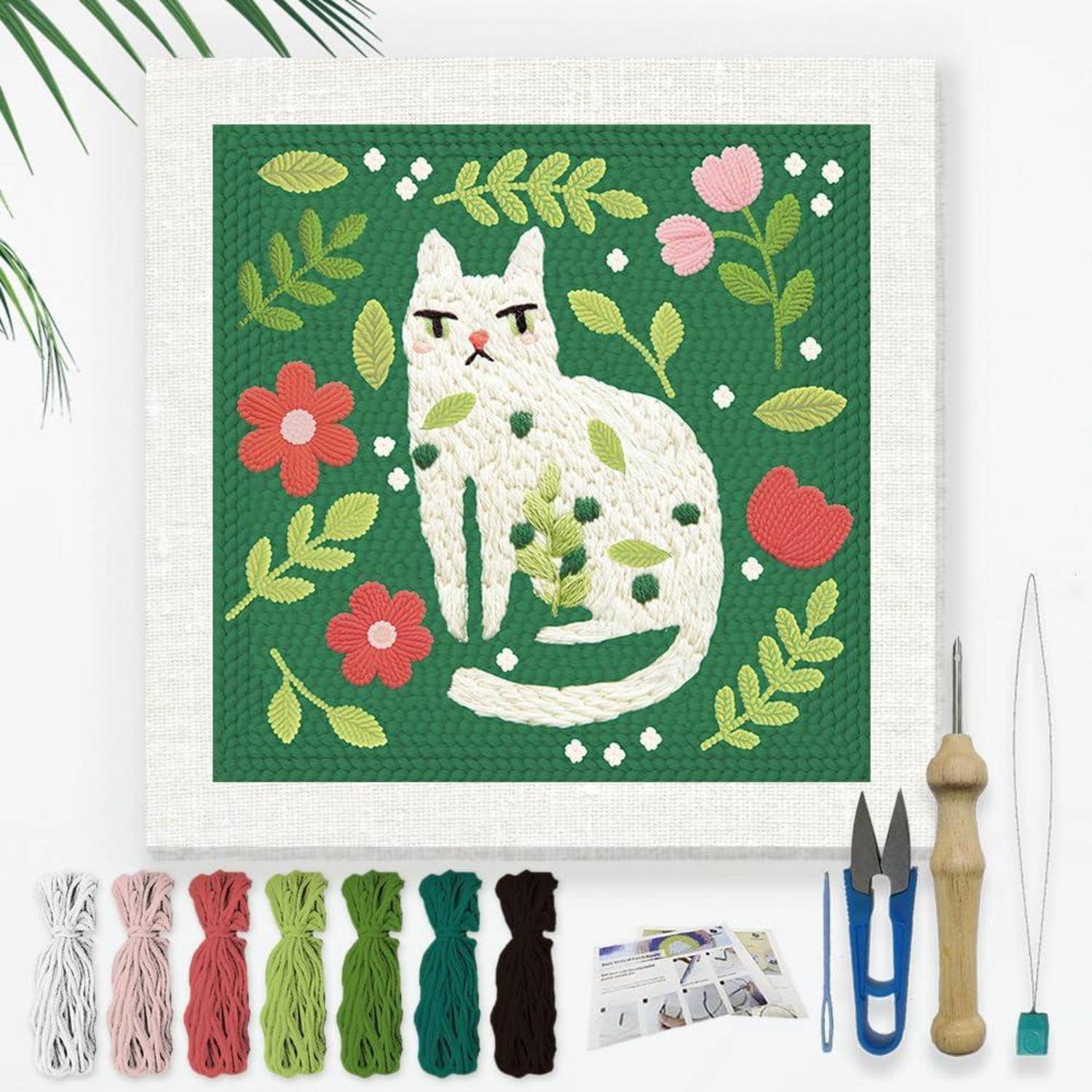 Stickset mit einer weißen Katze auf grünem Stoff mit Blumen und Blättern, mit Garn, Werkzeugen und Anleitung darunter angeordnet.