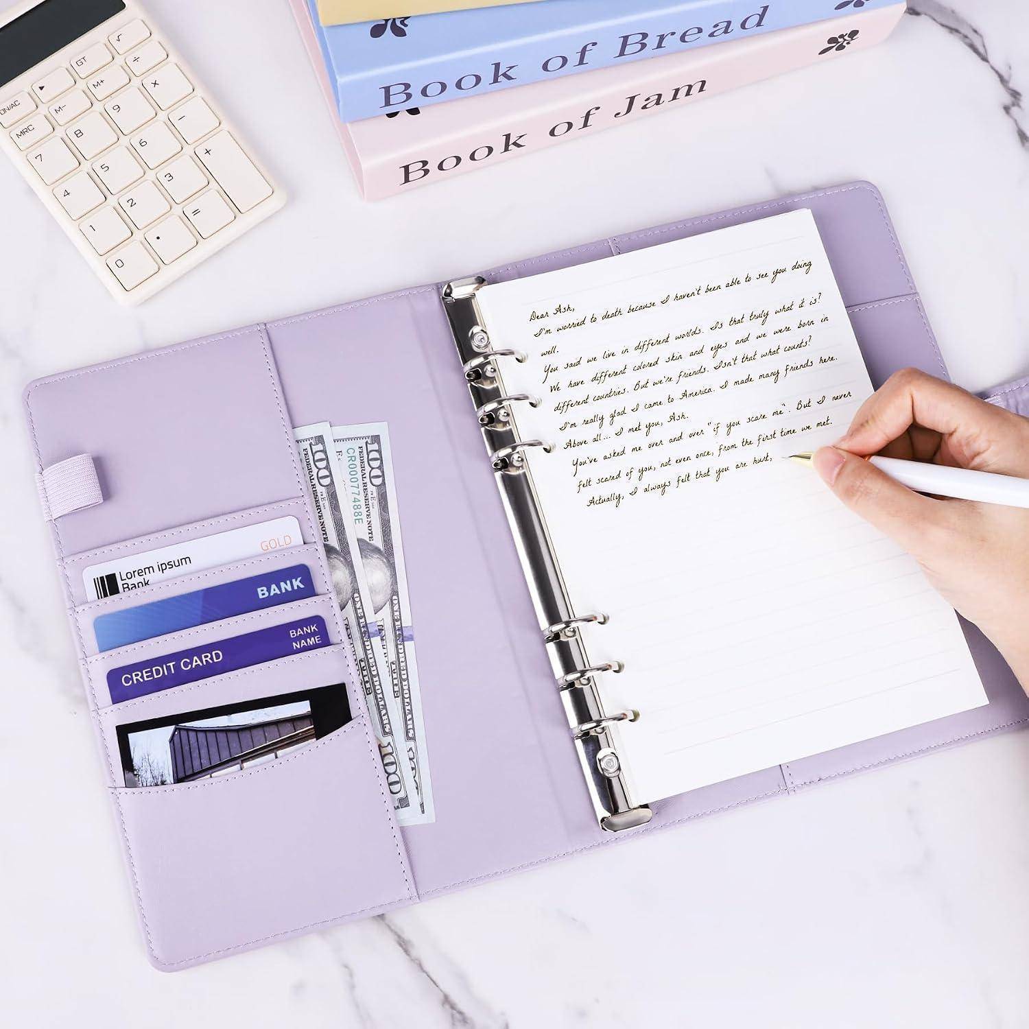 Offener lavendelfarbener Ringordner-Notizbuch mit Stift, der auf einer Seite schreibt, Bargeld und Karten in Taschen, neben einer Tastatur und beschrifteten Büchern auf einem Schreibtisch.
