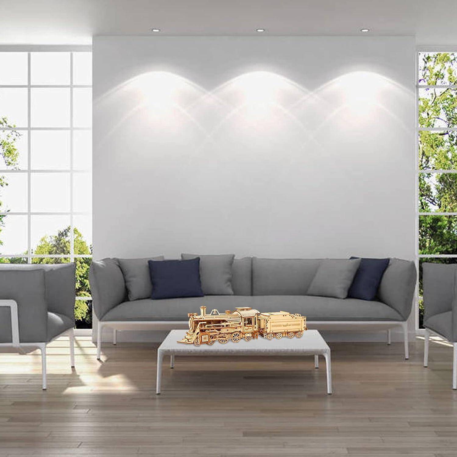 Modernes Wohnzimmer mit grauem Sofa und Stühlen um einen Couchtisch, auf dem eine hölzerne Spielzeugeisenbahn ausgestellt ist, beleuchtet durch Wandstrahler und große Fenster.