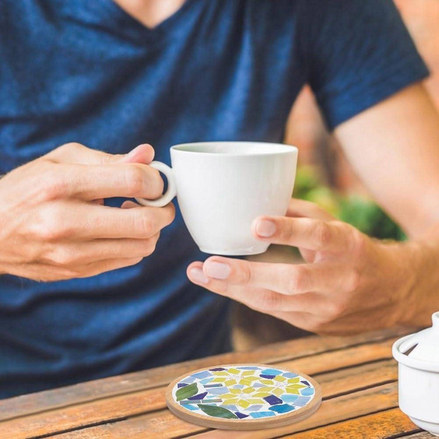 Eine Person in einem blauen Hemd hält eine weiße Tasse über einen Holztisch mit einem bunten Mosaikuntersetzer und einer teilweise sichtbaren Teekanne.