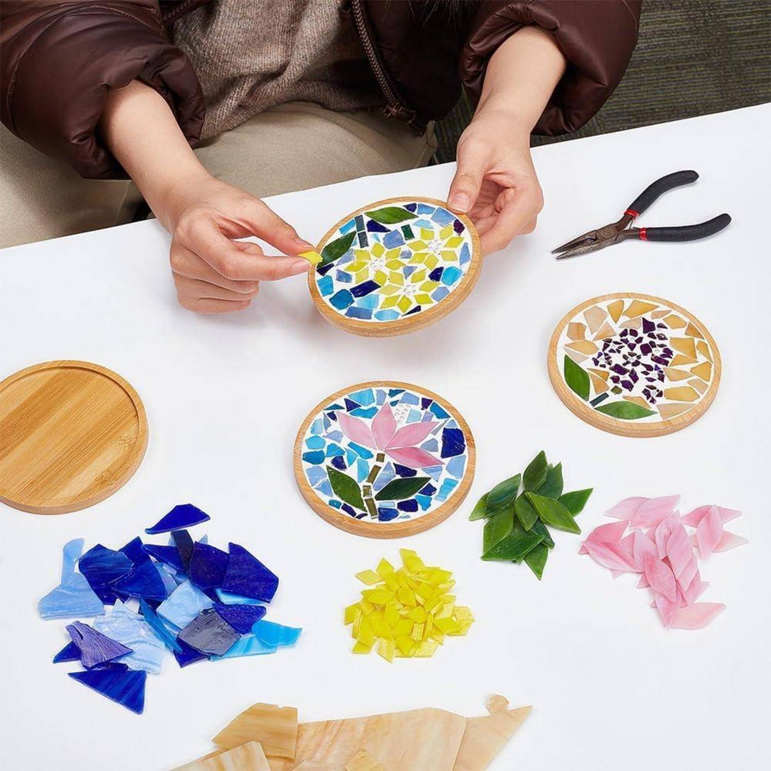 Person, die bunte Mosaik-Untersetzer auf einem weißen Tisch mit Glassplittern, Zangen und fertigen runden Designs zusammensetzt.