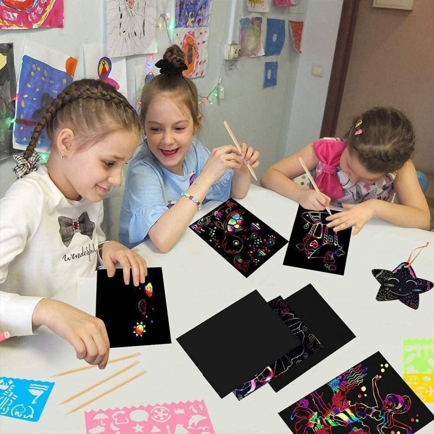 Drei junge Mädchen sitzen an einem Tisch und verwenden Holzstöcke, um bunte Designs auf schwarzen Kunstkarten in einer Kunstaktivität im Klassenzimmer zu kratzen.