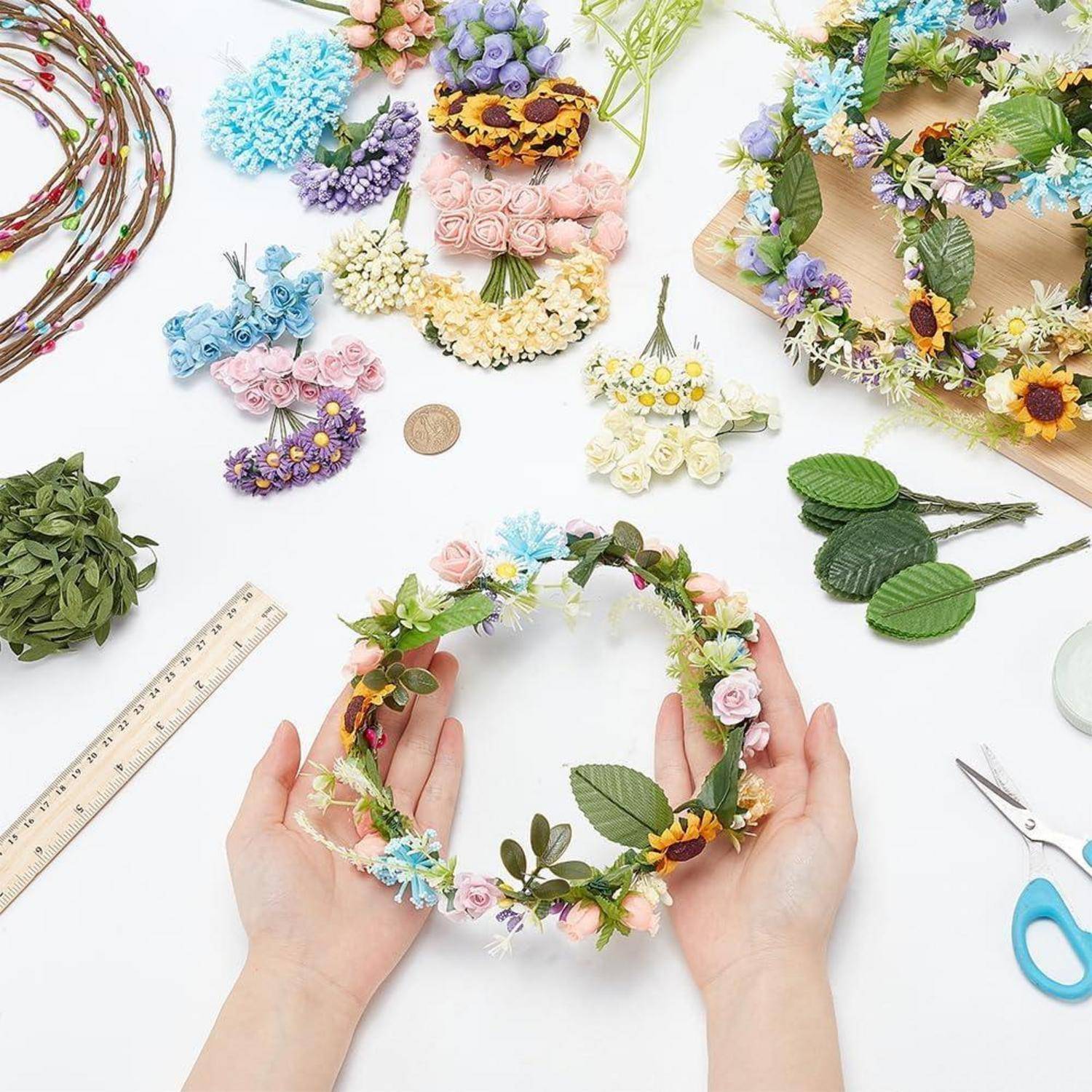 Hände halten einen floralen Kranzhandwerk-Ring mit kleinen Pastellblumen und Grünzeug auf einem weißen Tisch, umgeben von Bastelmaterialien und Blumenstücken.