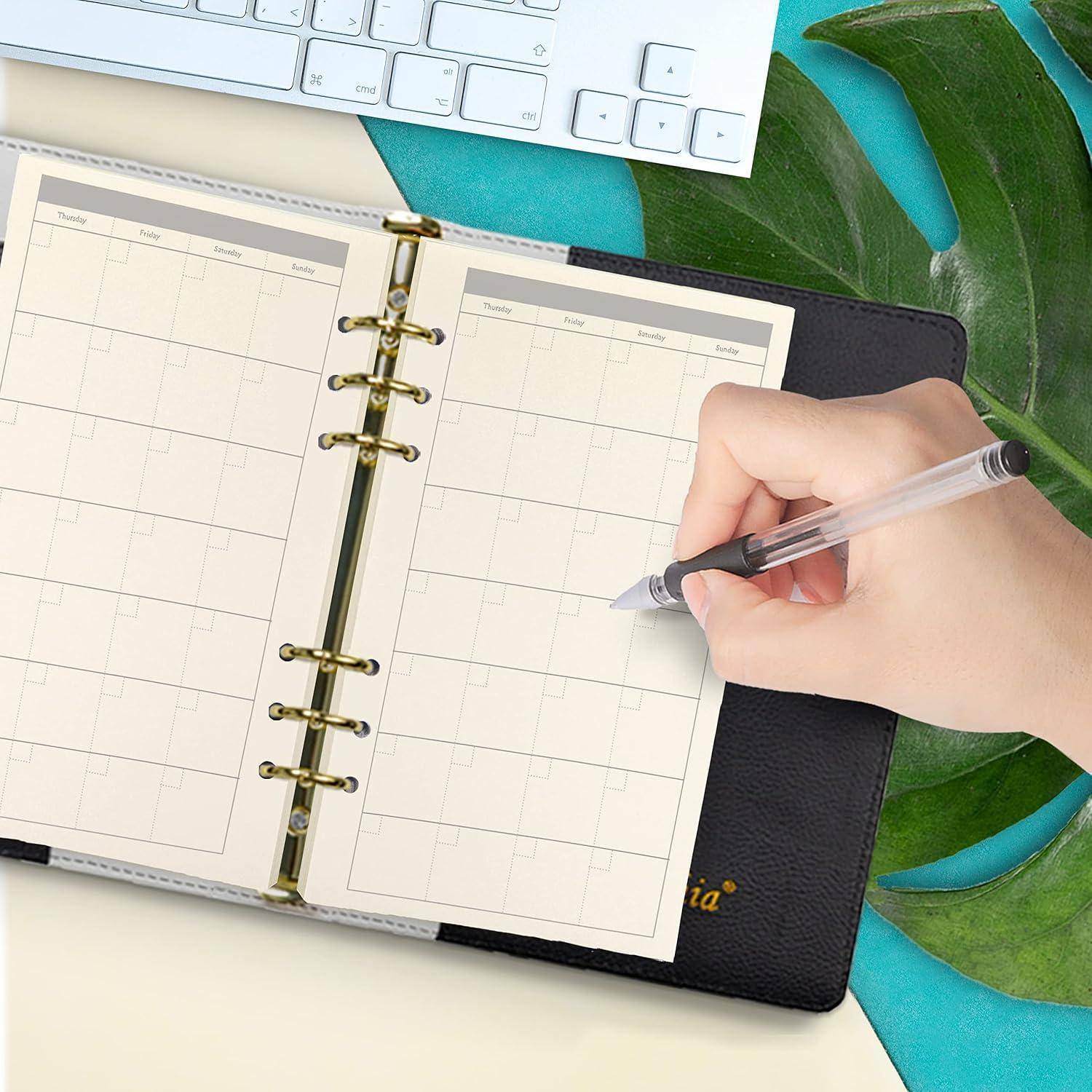 Eine Hand schreibt in einem spiralgebundenen Wochenplaner neben einer Computertastatur und einem grünen Blatt und deutet auf Terminplanung oder Planungsarbeit hin.