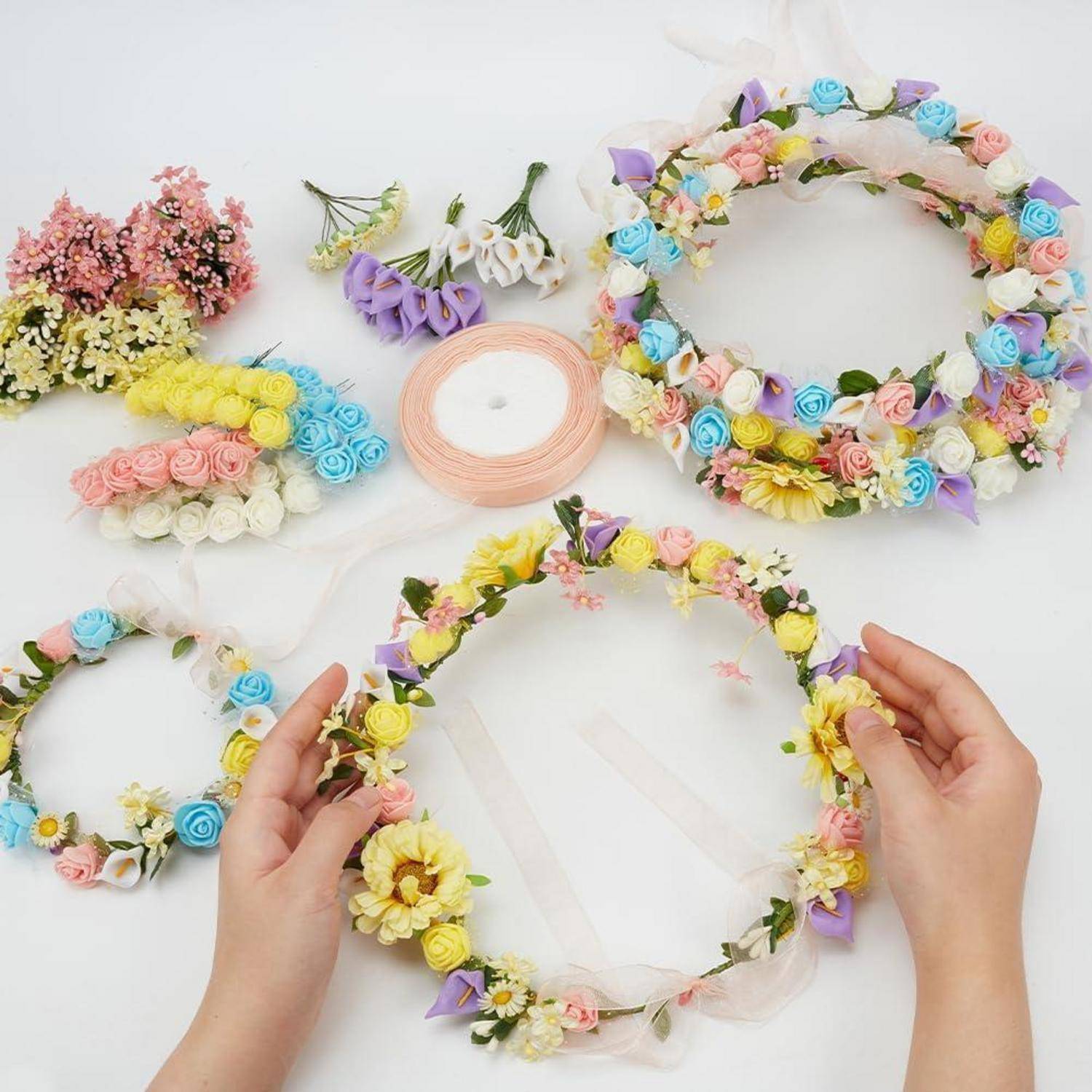 Hände, die eine Pastellblumenkrone auf einer weißen Oberfläche formen, mit mehreren fertiggestellten Blumenkränzen und lockeren künstlichen Blumen in der Nähe.