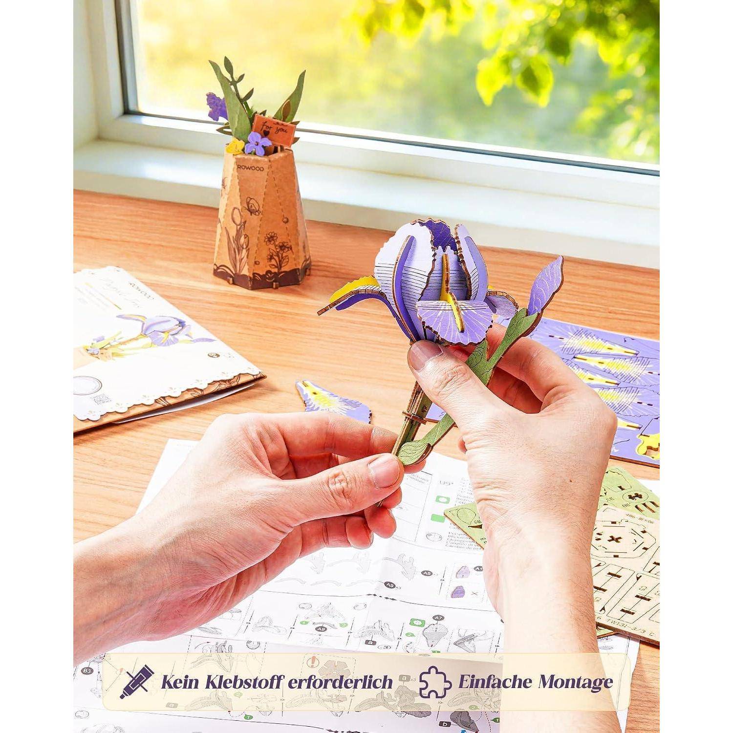 Hände, die ein lila Papierblumenbastelprojekt auf einem Tisch zusammensetzen, mit einer Verpackung, auf der „Kein Klebstoff erforderlich