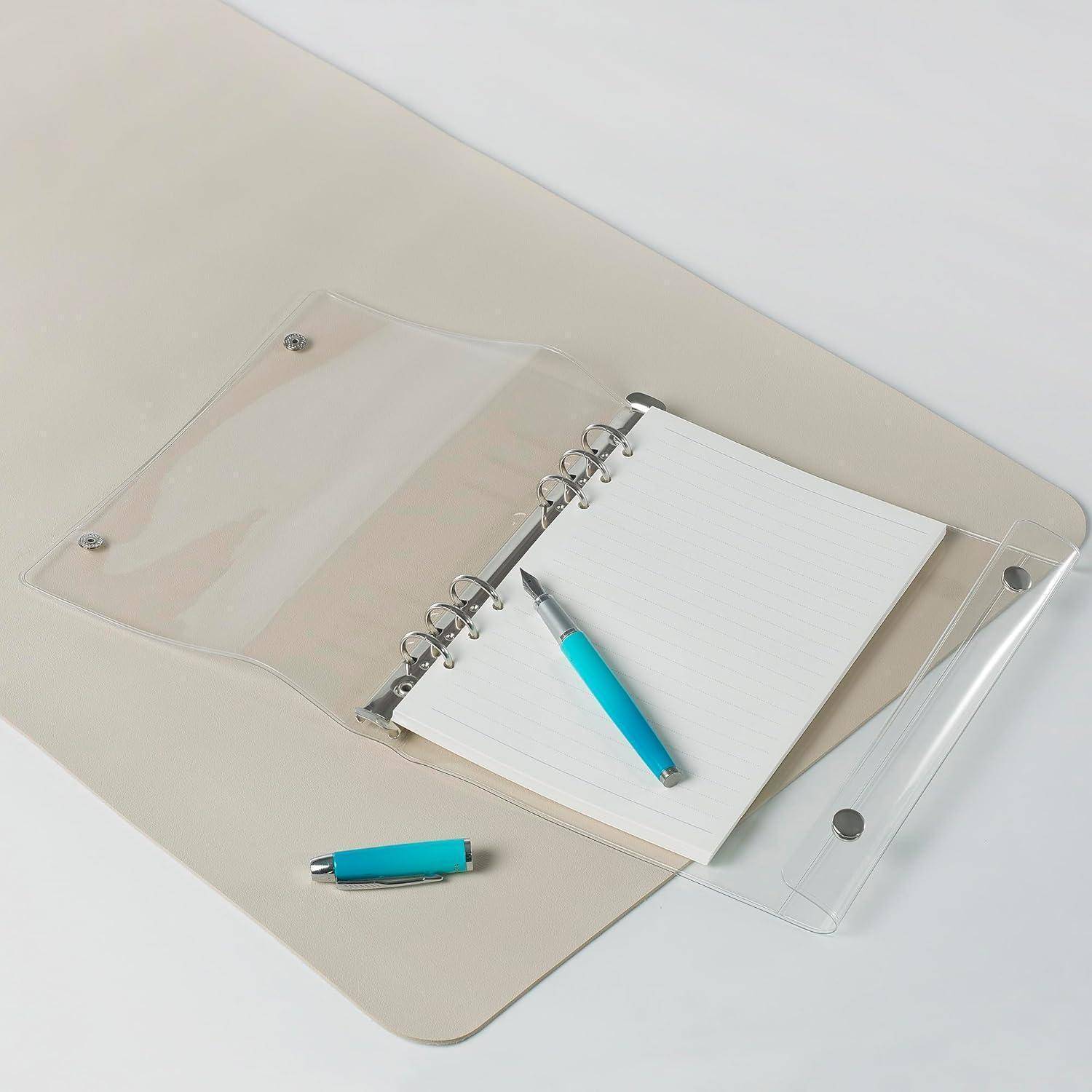 Transparenter Kunststoffordner mit einem kleinen Ringnotizbuch und zwei türkisfarbenen Stiften auf einer beigefarbenen Schreibunterlage vor weißem Hintergrund.