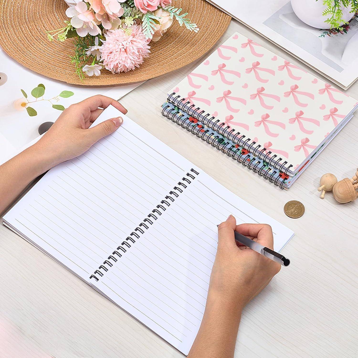 Hände, die in einem offenen linierten Notizbuch auf einem ordentlichen Schreibtisch schreiben, mit einem anderen floralen Spiralnotizbuch, Blumen und einem Stift in der Nähe.