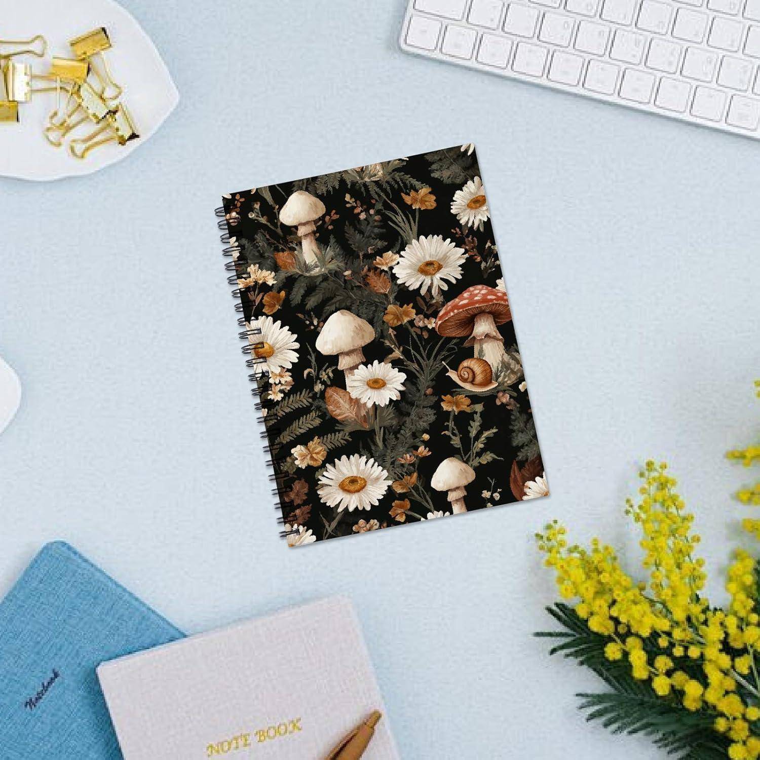 Ein Notizbuch mit dunklem Blumenpilz-Cover liegt auf einem Schreibtisch neben einem blauen Notizbuch, einer Tastatur, Blumen und Büroklammern.
