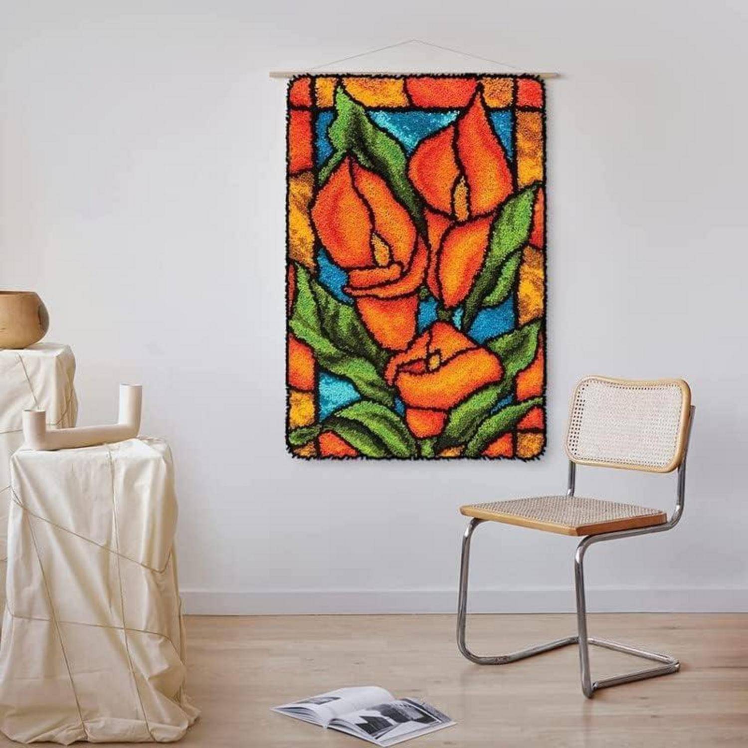 Farbenfrohe Blumen-Wandteppich mit orangefarbenen Blüten und grünen Blättern, der an einer weißen Wand in einem minimalistischen Raum hängt, neben einem Korbstuhl und einem drapierten Tisch.