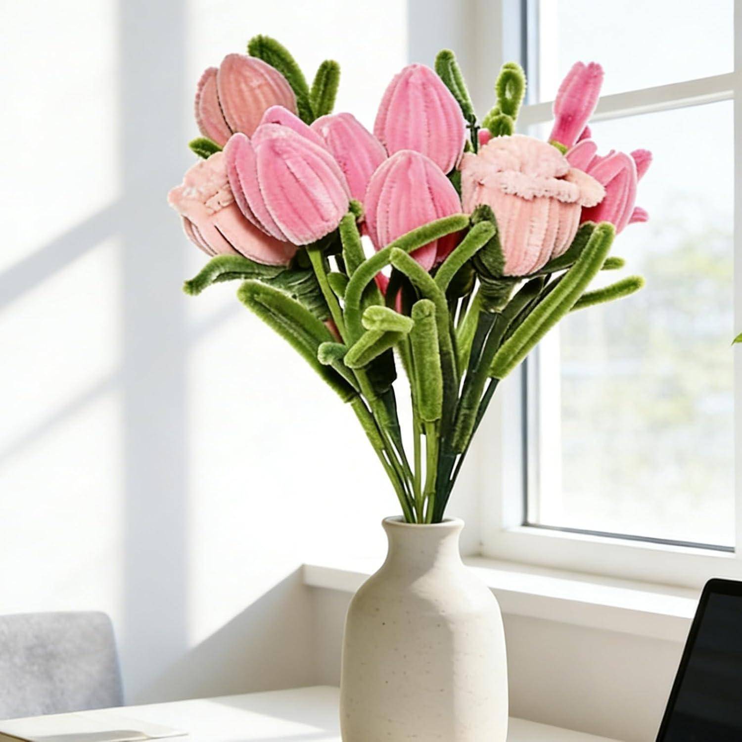 Rosa künstlicher Blumenstrauß in einer weißen Vase auf einem sonnigen Schreibtisch neben einem Fenster, wobei ein Teil eines Laptops sichtbar ist.