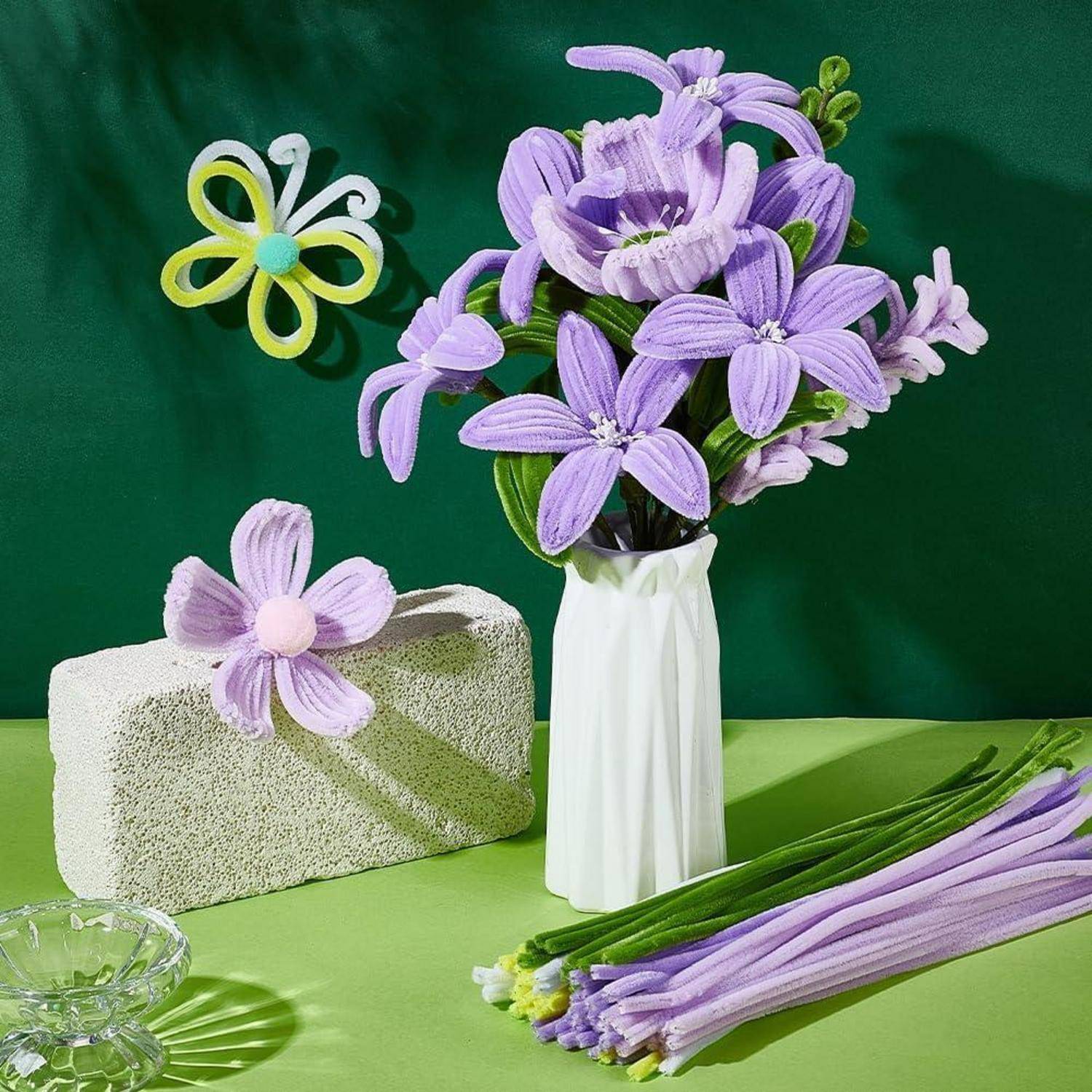 Strauß aus violetten handgefertigten Blumen in einer weißen Vase, mit passenden Blumenelementen und grünen Stielen auf einem Tisch vor grünem Hintergrund arrangiert.