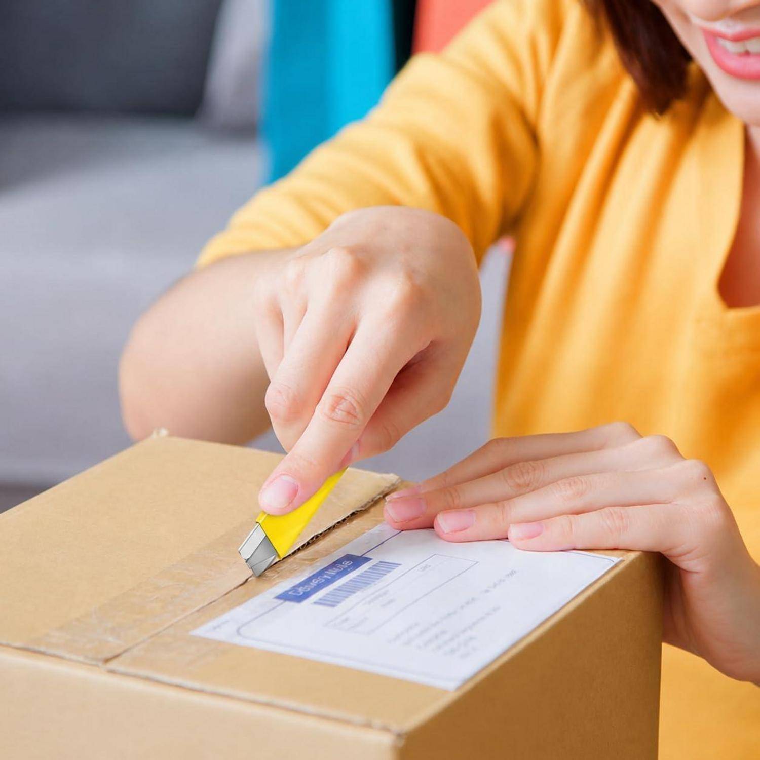 Person in einem gelben Hemd, der ein Teppichmesser benutzt, um ein versiegeltes Kartonpaket mit einem Versandetikett zu öffnen.