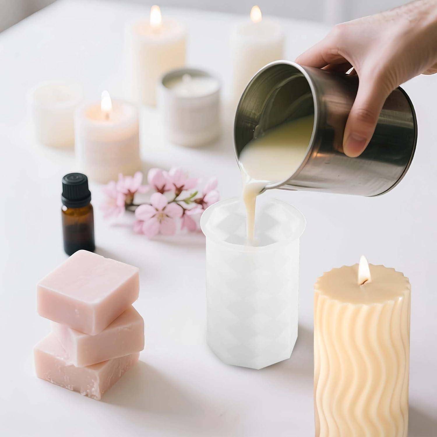 Hand gießt geschmolzenes Wachs aus einem Metallkrug in eine weiße Kerzenform, mit fertigen Kerzen, Wachsblöcken, Blumen und einer Duftflasche auf einem Tisch.