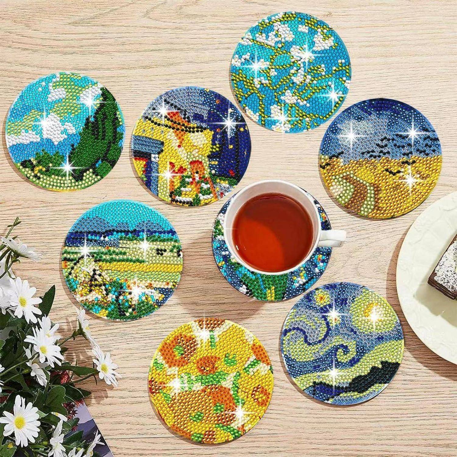 Bunte Perlenuntersetzer, die um eine Tasse Tee auf einem Holztisch angeordnet sind, wobei jeder Untersetzer ein anderes Landschafts- oder Blumenmosaik-Design zeigt.
