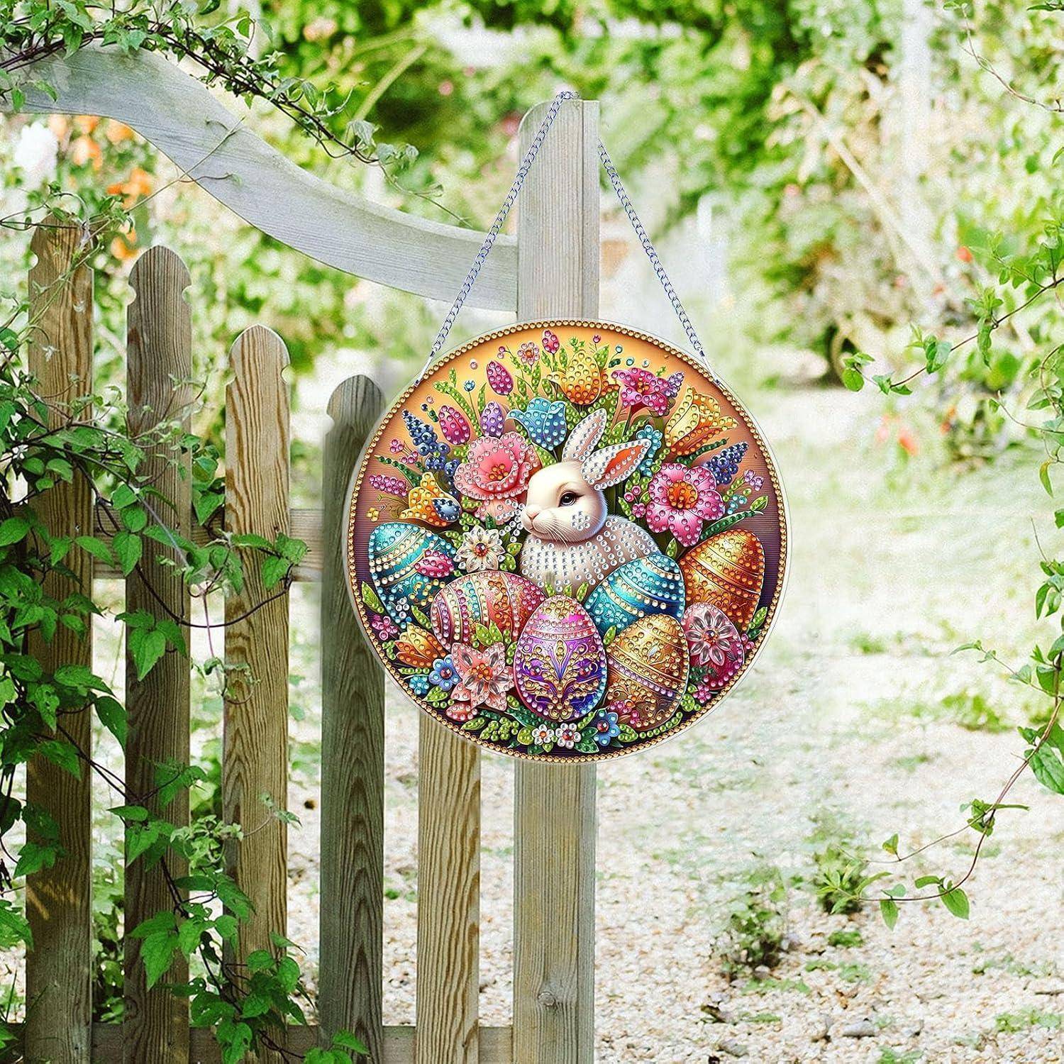 Rundes florales Osterei-Ornament mit Hasengeficht hängt an einem Gartenzaun, umgeben von Grünflächen und einem Kiesweg.