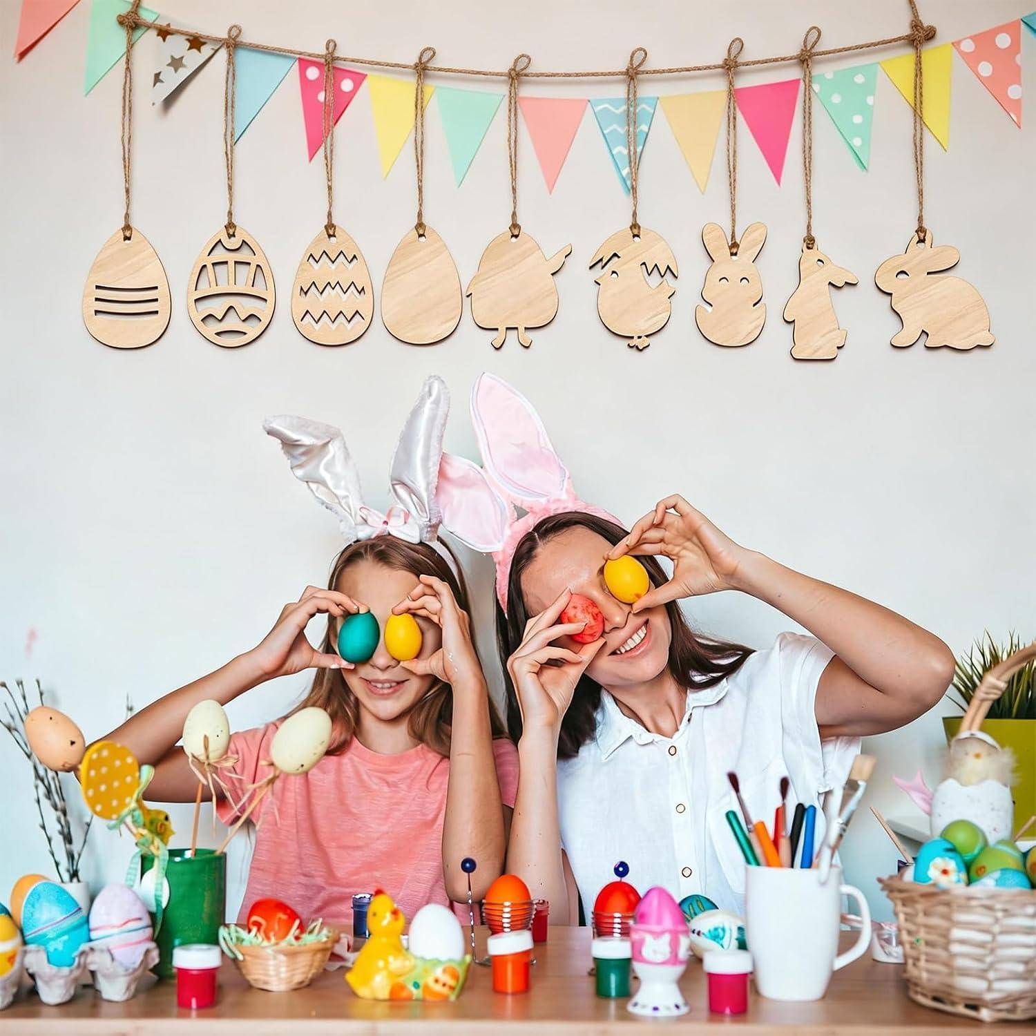 Zwei Mädchen mit Hasenohren halten bunte Eier über ihre Augen an einem Oster-Basteltisch mit bemalten Eiern, Körben und Dekorationen.