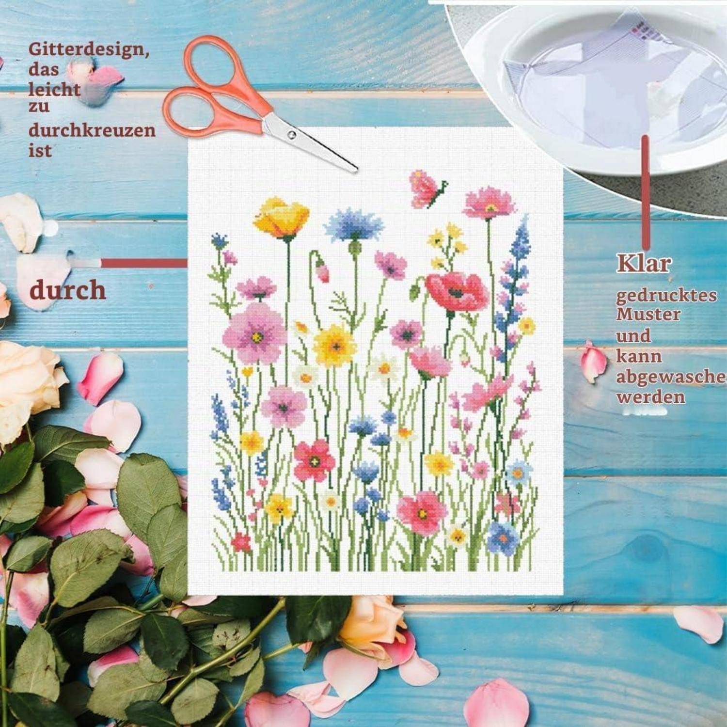 Stickereimuster auf weißem Stoff mit bunten Wildblumen, mit Schere und Stickrahmen auf einem blauen Holztisch; deutscher Text zeigt an, dass das Design einfach zu kreuzsticken und waschbar ist.