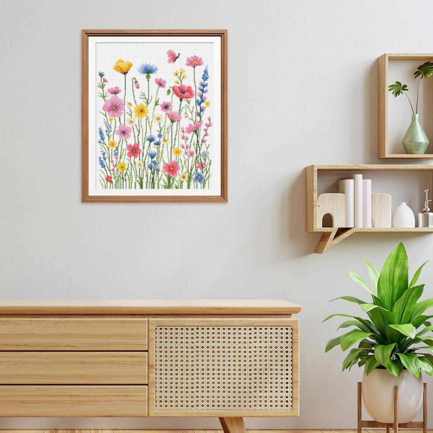 Gerahmtes Aquarell mit bunten Wildblumen an einer hellen Wand über einem Holzschrank mit Regalen, einer Vase und einer Topfpflanze in einem minimalistischen Zimmer.