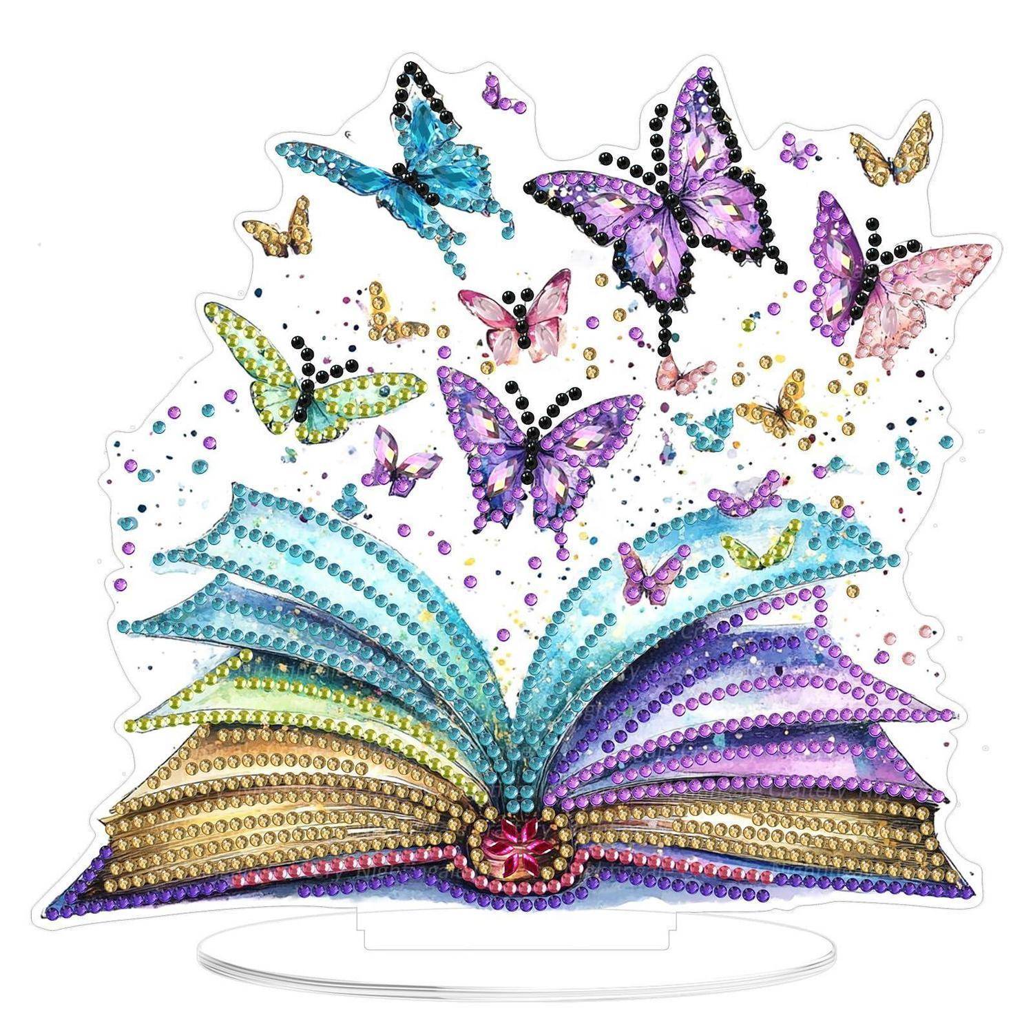 Ein aufgeschlagenes Buch mit regenbogenfarbenen Seiten und funkelnden Schmetterlingen, die nach oben ausbrechen, eine dekorative Diamantkunst-Stilanzeige auf einem transparenten Ständer.