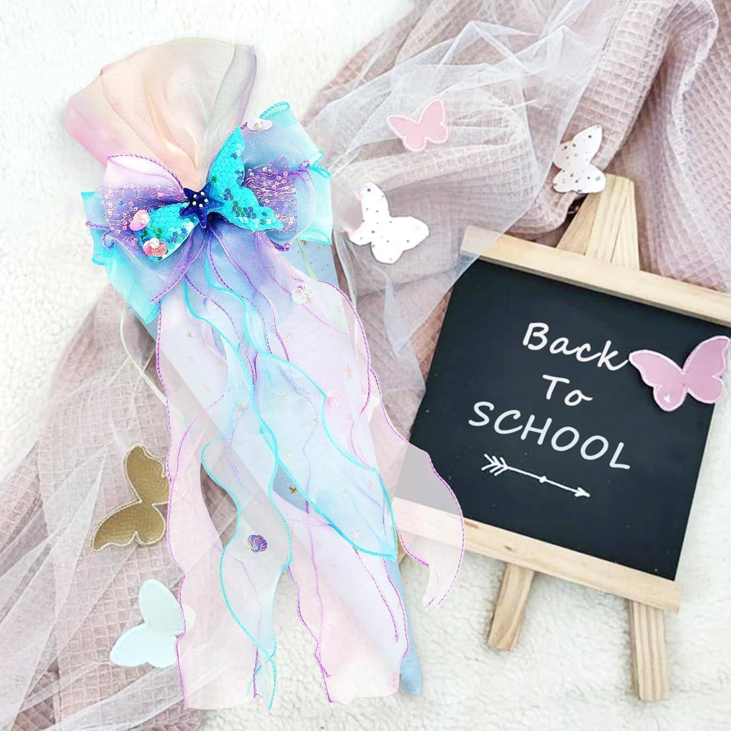 Dekoriertes Pastell-Zurück-zur-Schule-Schild auf einem kleinen Ständer mit Schmetterlingen, neben einem durchsichtigen Zauberstab mit Bändern oder Geschenkfavor in zartem Rosa, Blau und Lavendel.