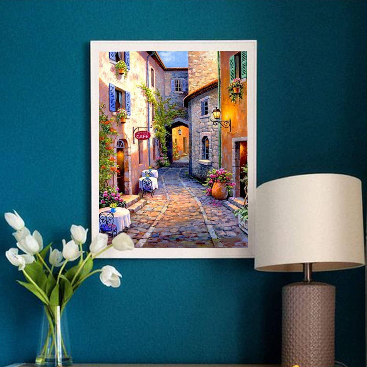 Gerahmtes Gemälde einer engen Kopfsteinpflaster-Gasse mit Café-Tischen und Blumen an einer blaugrünen Wand, flankiert von einer Vase mit weißen Tulpen und einer Tischlampe.