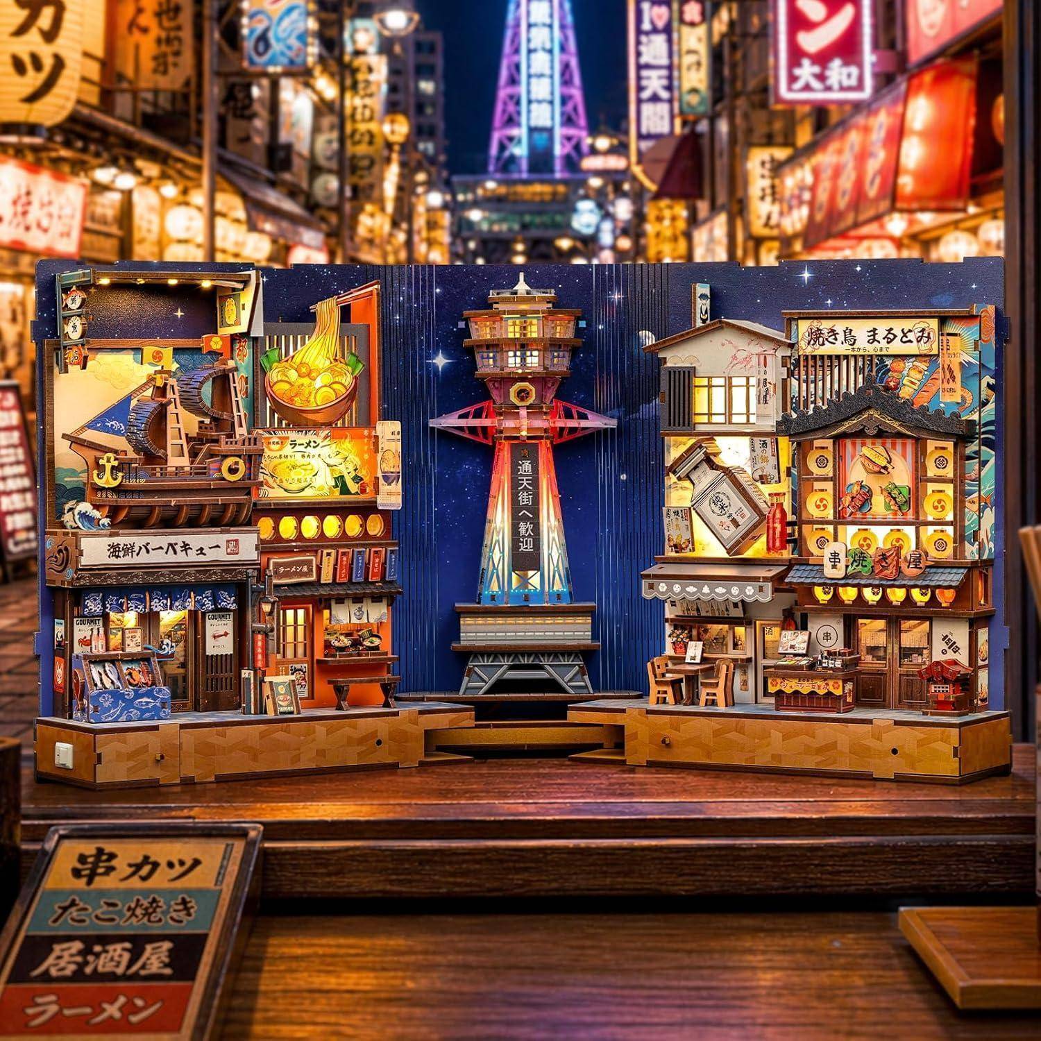 Miniatur-Japanischer Straßenmarkt mit leuchtenden Ladenfront und einem zentralen Turm vor nächtlicher Stadtkulisse.