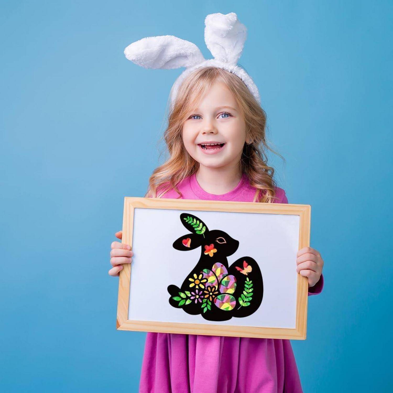 Lächelndes Mädchen mit Hasenohren hält eine gerahmte schwarze Hasenschablone, die mit bunten Blumen verziert ist, vor einem blauen Hintergrund.