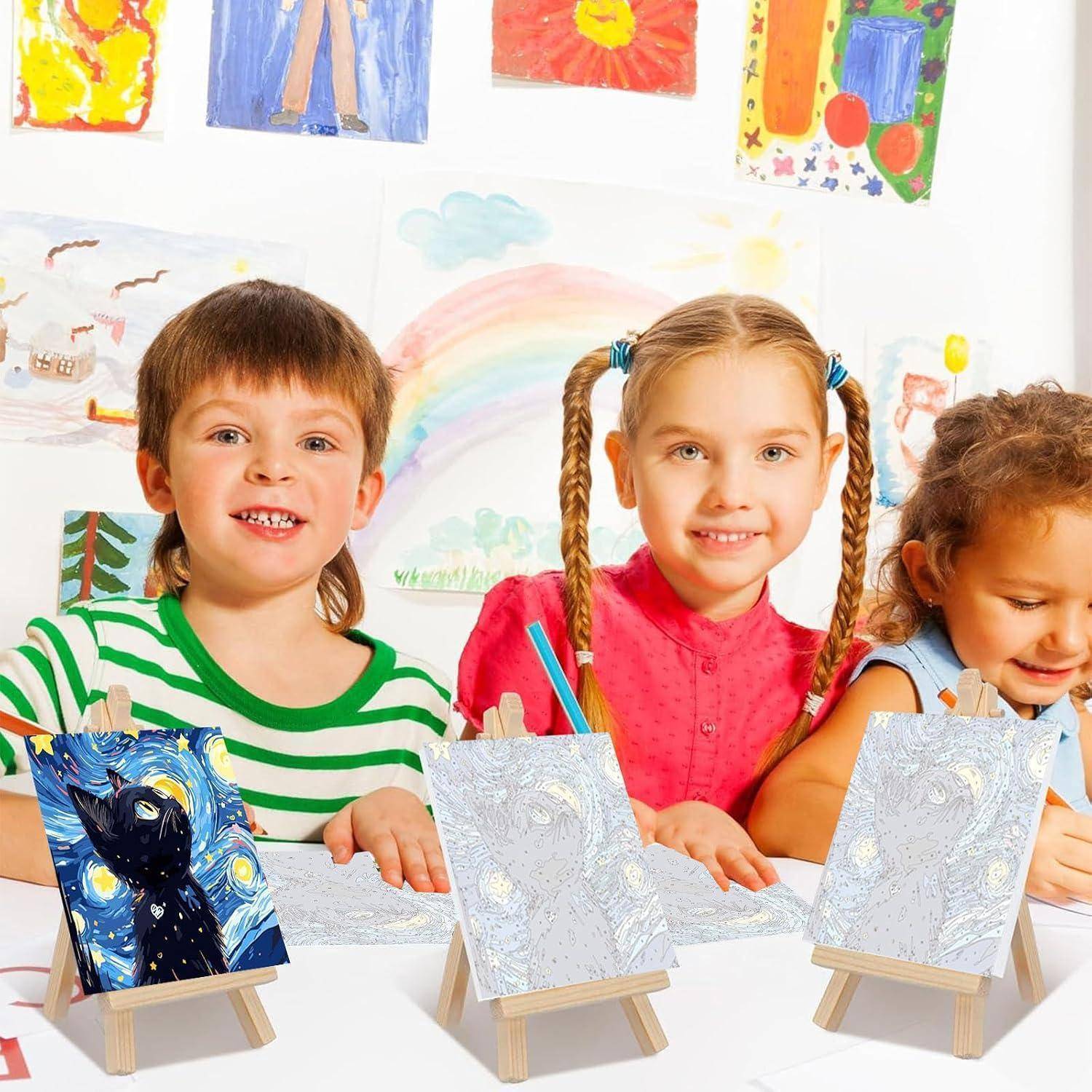 Drei kleine Kinder sitzen an einem Tisch in einem Kunstklassenzimmer, lächelnd neben kleinen Staffeleimalereien und ihren bunten Zeichnungen an der Wand.