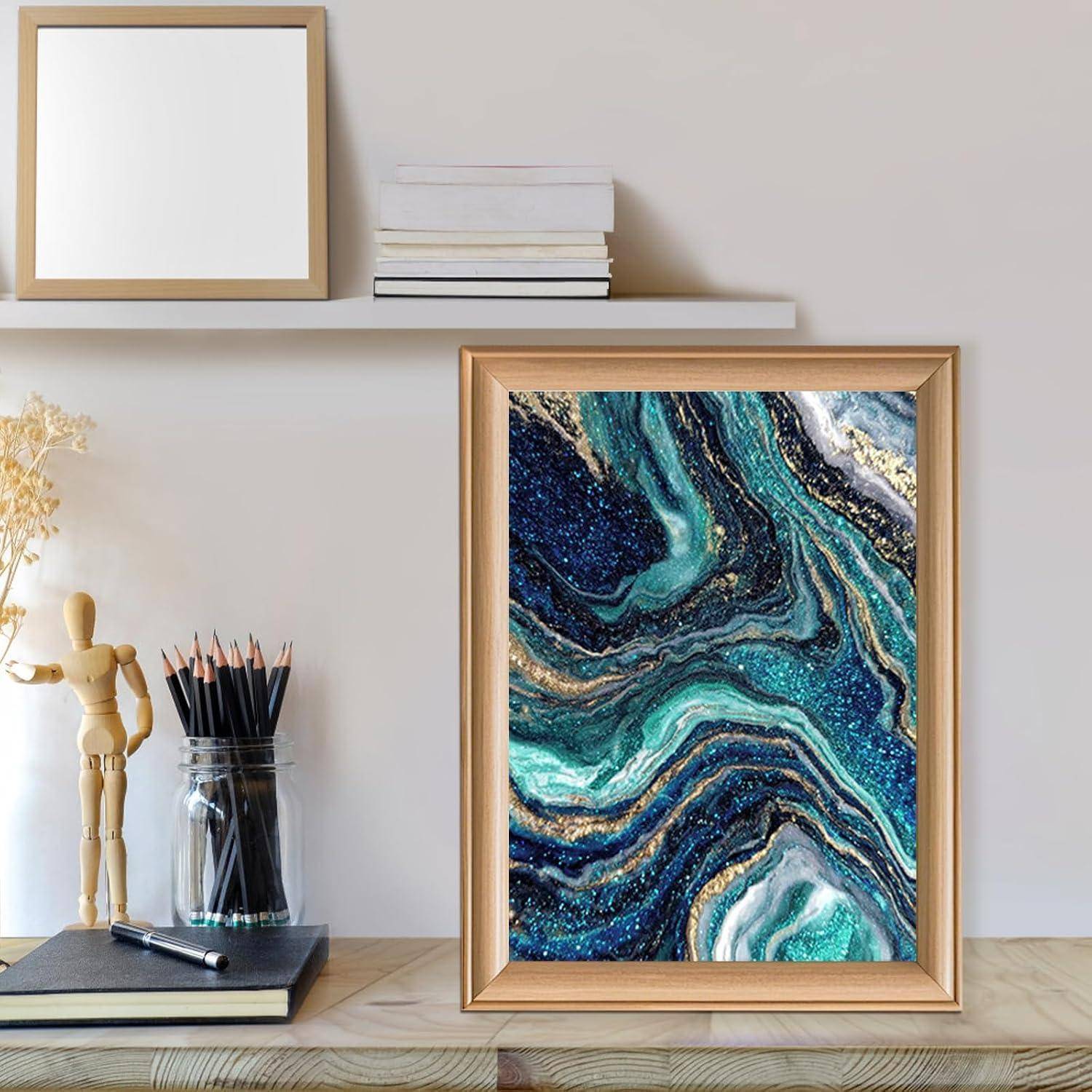 Gerahmtes abstraktes Kunstwerk mit türkisfarbenem, marineblauem und goldenem Fluid-Marbling sitzt auf einem Holzschreibtisch neben Bleistiften, einer Gliederpuppe und Büchern auf einem Regal.