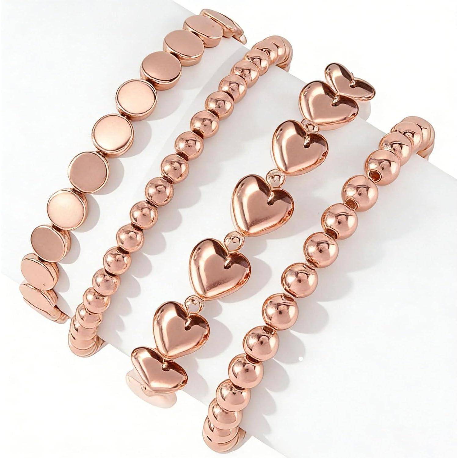 Vier rosegoldfarbene Armbänder auf einer weißen Oberfläche angeordnet: zwei mit runden Perlen, eines mit flachen Scheiben und eines mit herzförmigen Anhängern.