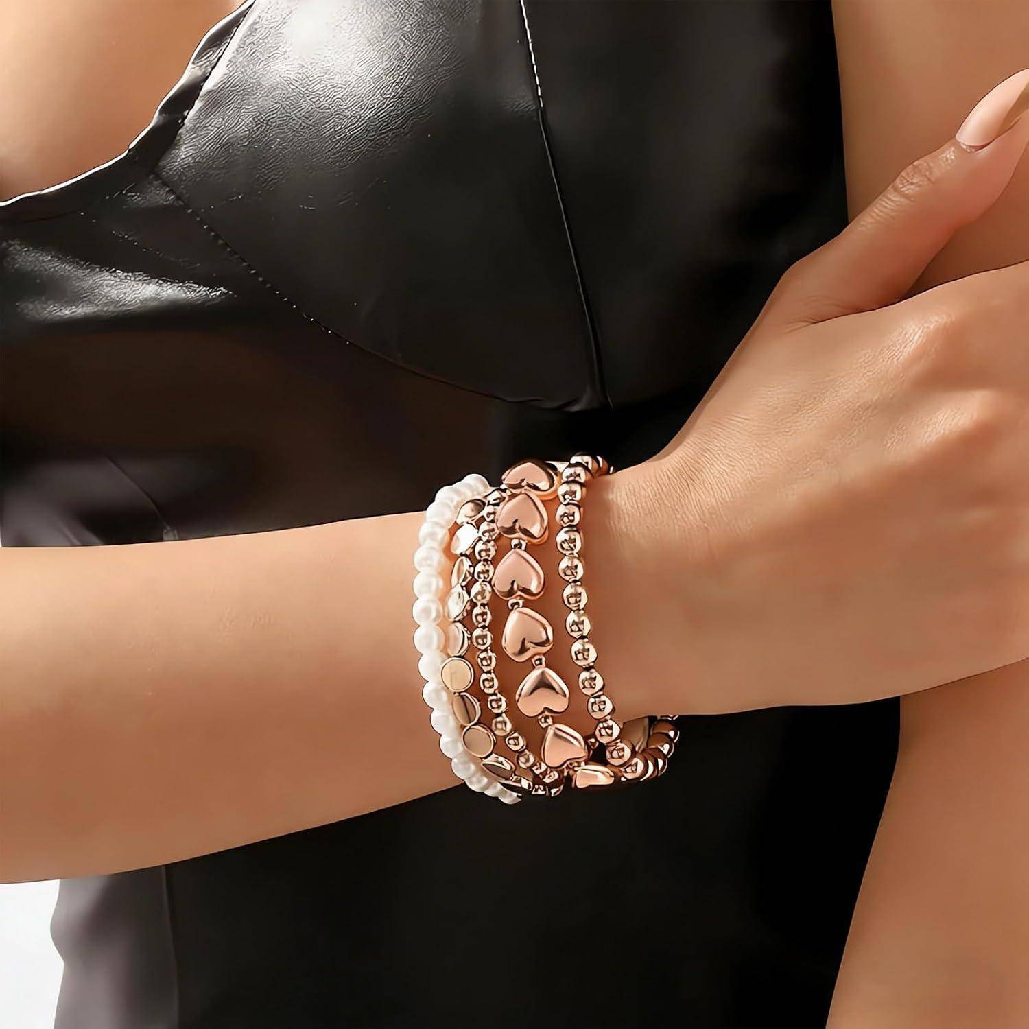 Nahaufnahme eines Handgelenks mit gestapelten Armbändern mit rosegoldfarbenen Herzanhängern und weißen Perlen vor einem schwarzen Outfit.