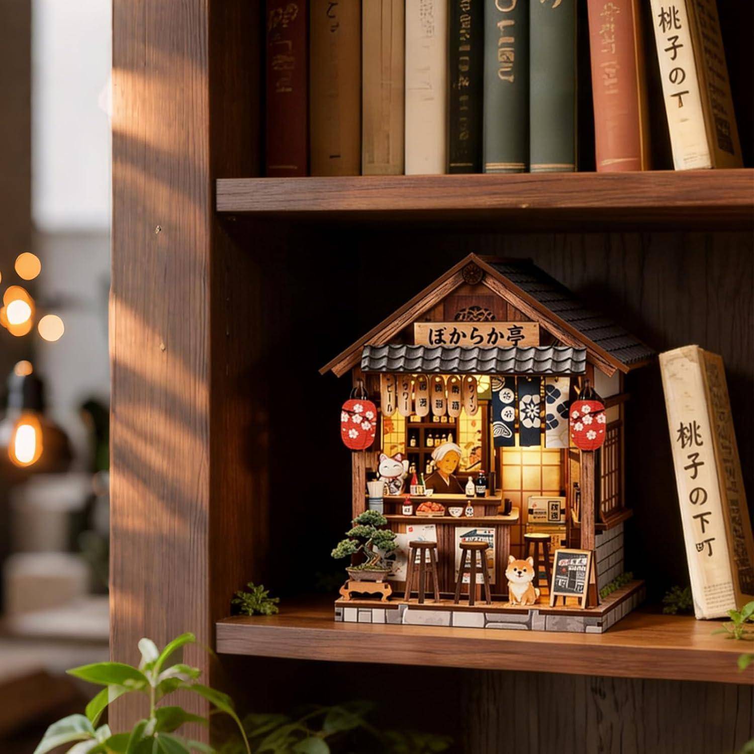 Miniatur-Japanisches Ladengeschäft-Modell mit Laternen auf einem Bücherregal, umgeben von Büchern und Pflanzen in einer warmen Inneneinrichtung.