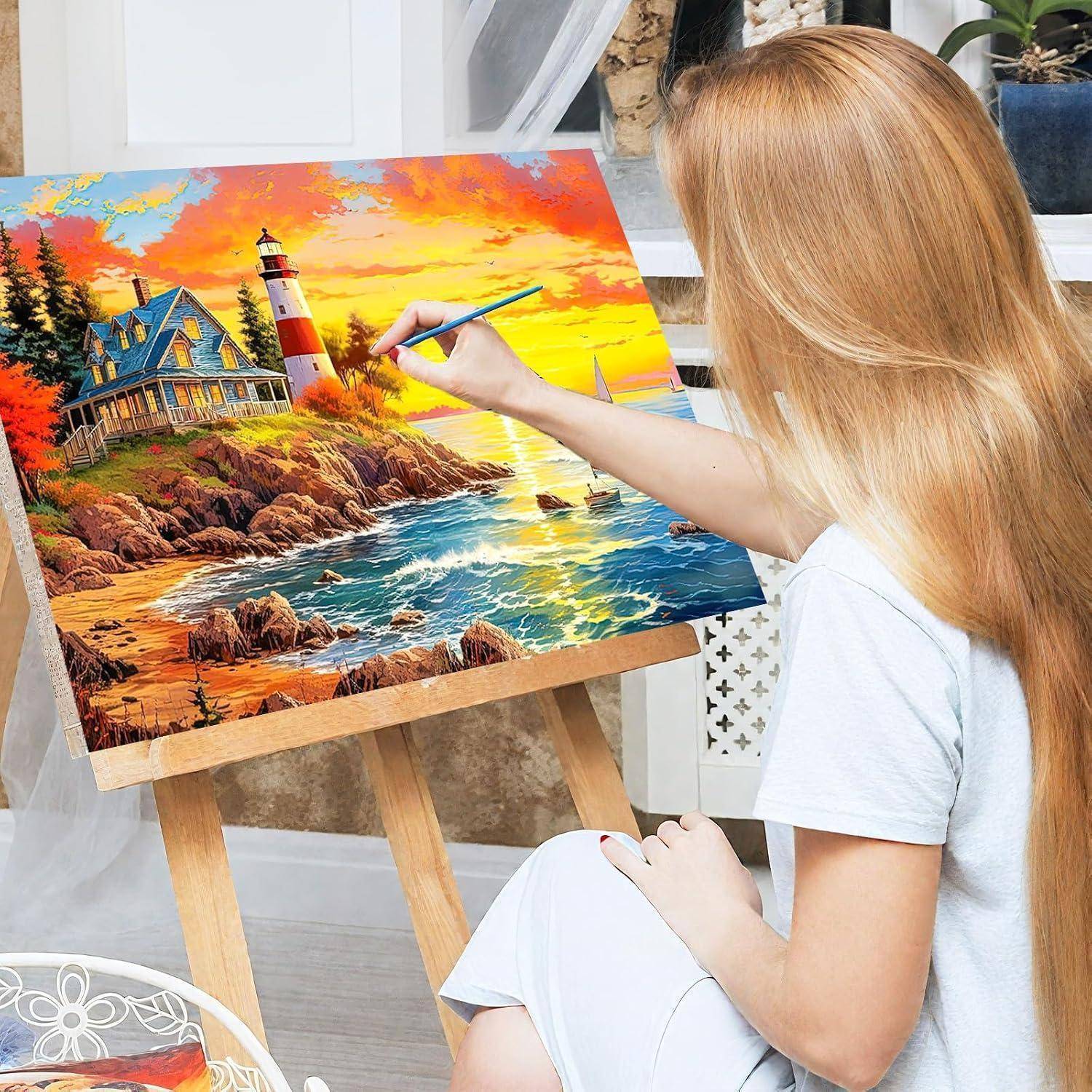 Eine Frau malt eine farbenfrohe Küstenszene auf eine Leinwand, die einen Leuchtturm, ein Haus, eine felsige Küste und einen Sonnenuntergangshimmel darstellt.