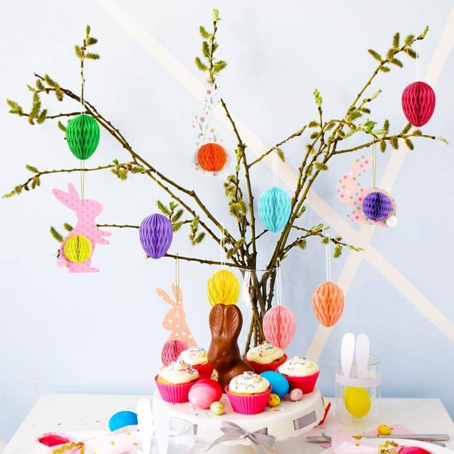 Ostergebäck-Präsentation mit Cupcakes, bunten Süßigkeitseiern und einem Schokoladenhäschen unter dekorierten Zweigen mit hängenden Papierei-Ornamenten.
