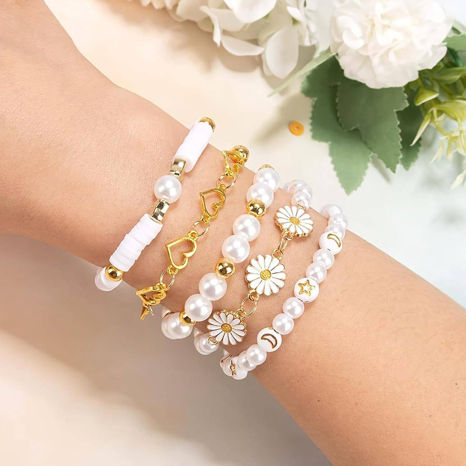 Nahaufnahme eines Handgelenks mit fünf übereinander gestapelten Armbändern mit weißen Perlen, goldenen Herzgliedern und Gänseblümchen-Anhängern auf einem weichen Blumenhintergrund.