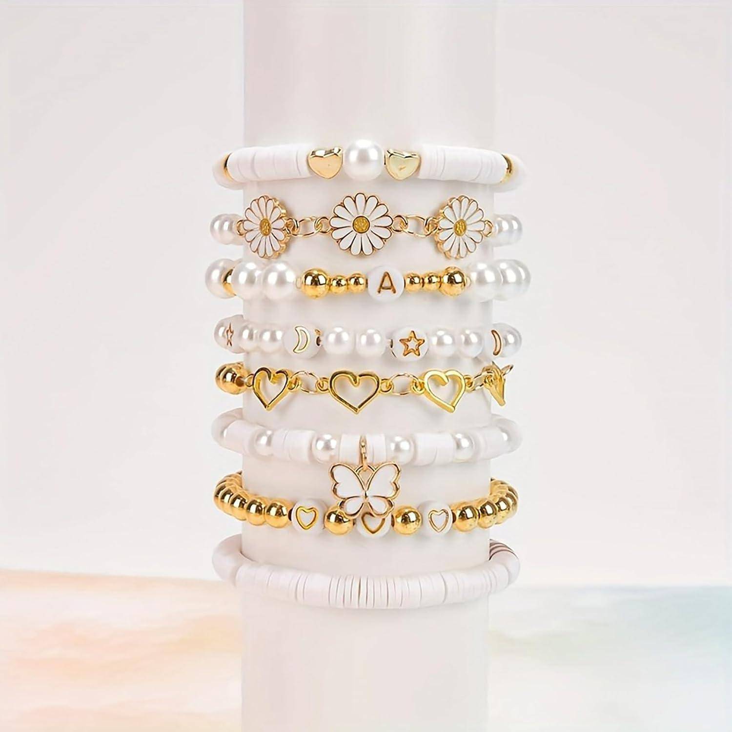 Stapel aus weißen und goldenen Perlmuttarmbändern mit Blumen-, Herz-, Perlen-, Stern-, Schmetterlings- und Initialen-Anhängern, ausgestellt auf einem weißen Ständer.