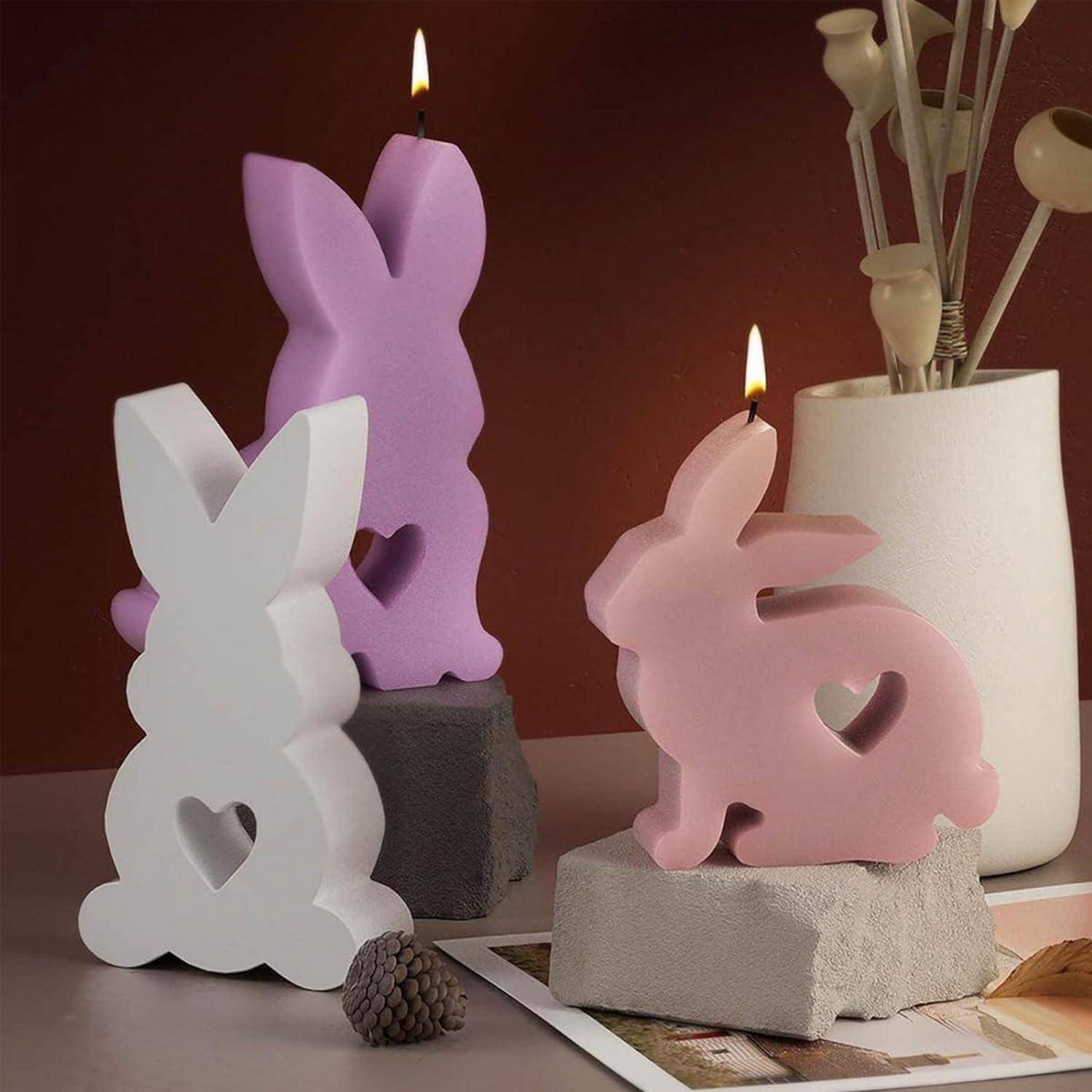 Dekorative osterhashenförmige Kerzen in Weiß, Lila und Rosa, die auf Steinblöcken neben einer weißen Vase mit getrockneten Stielen brennen.
