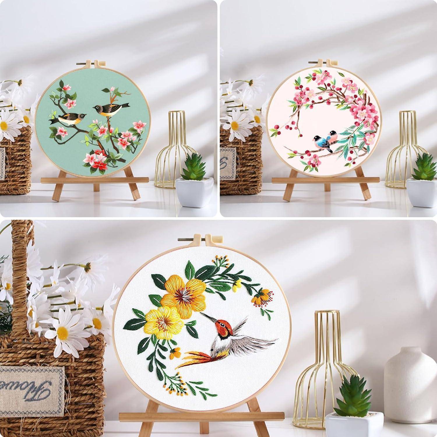 Drei bestickte Reifen-Kunstdesigns zur Schau gestellt: Vögel mit Blumen in Minz-, Rosa- und weißen Blumenkränzen sowie ein Kolibri inmitten gelber Blüten auf einem Holzständer.